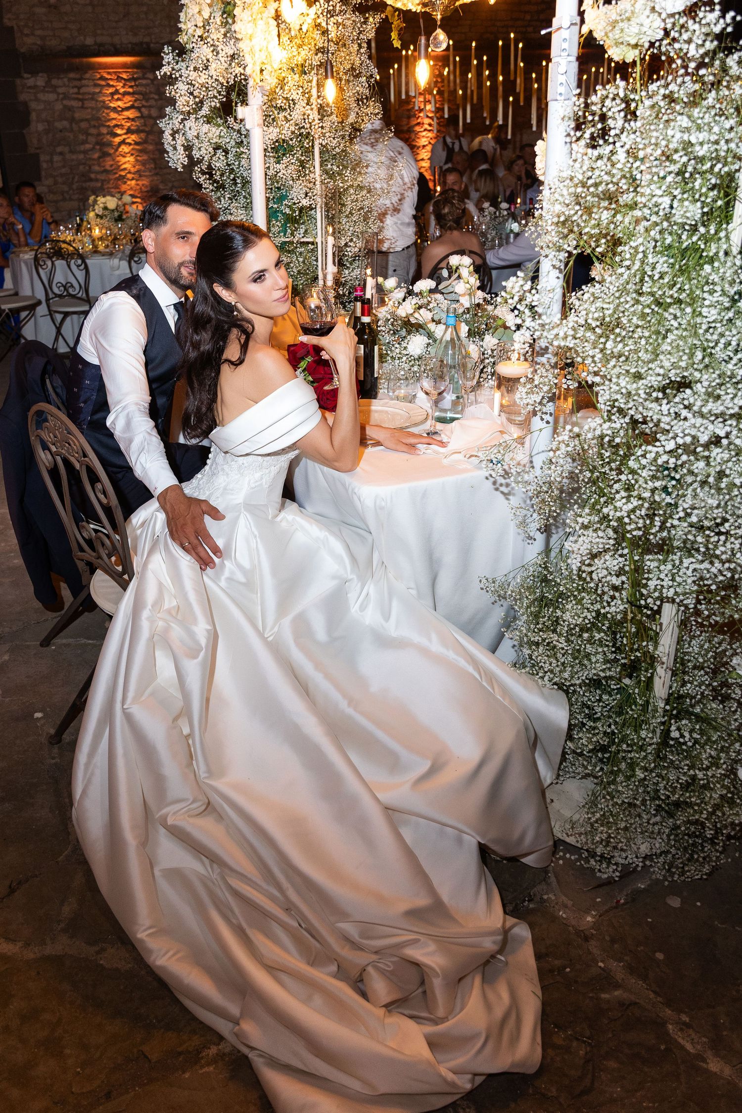 Romantic couple in white wedding attire dancing in a rustic venue decorated with delicate baby's breath flowers.
