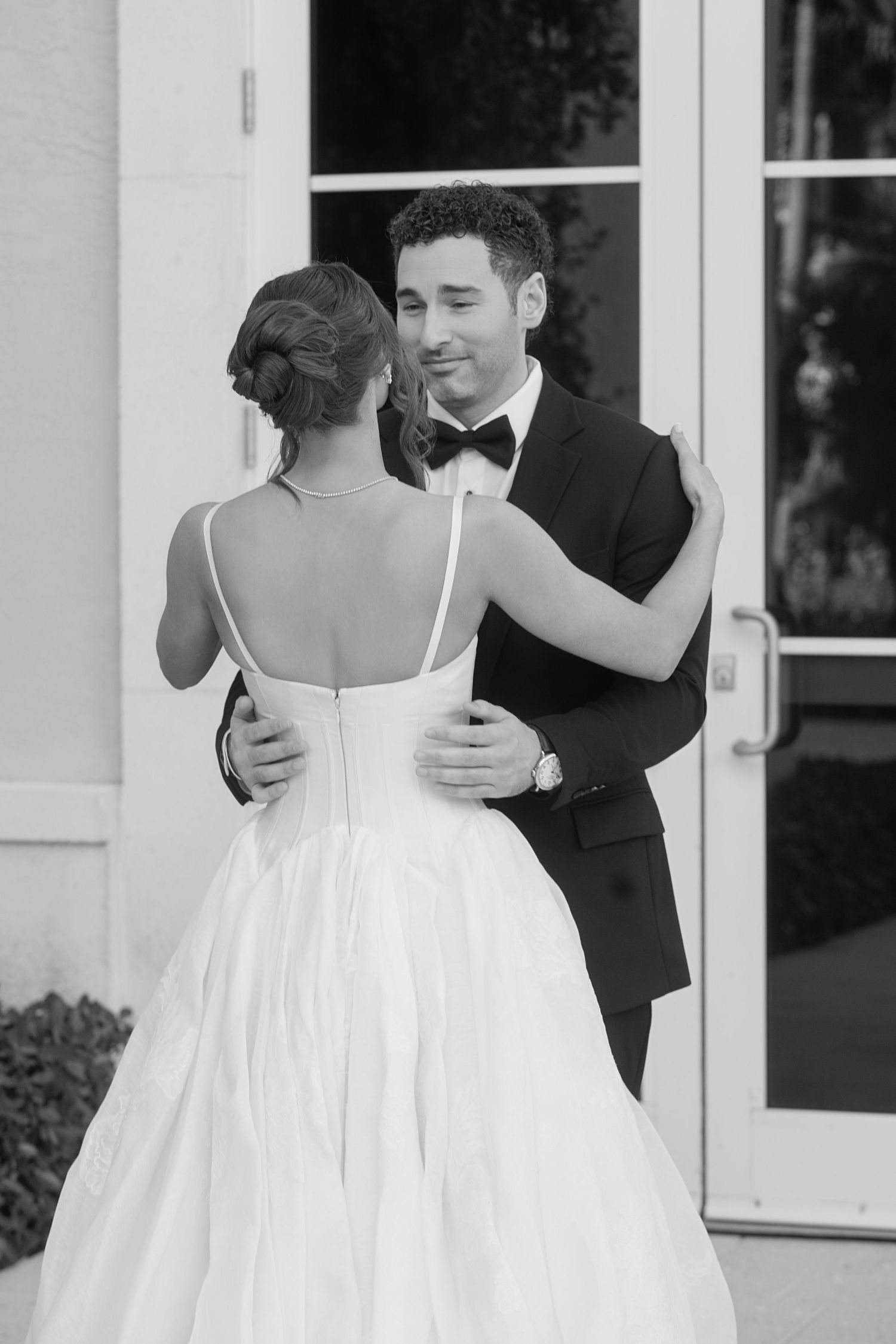 Black and white photo of a bride and groom sharing a first look, embracing outside elegant white doors.