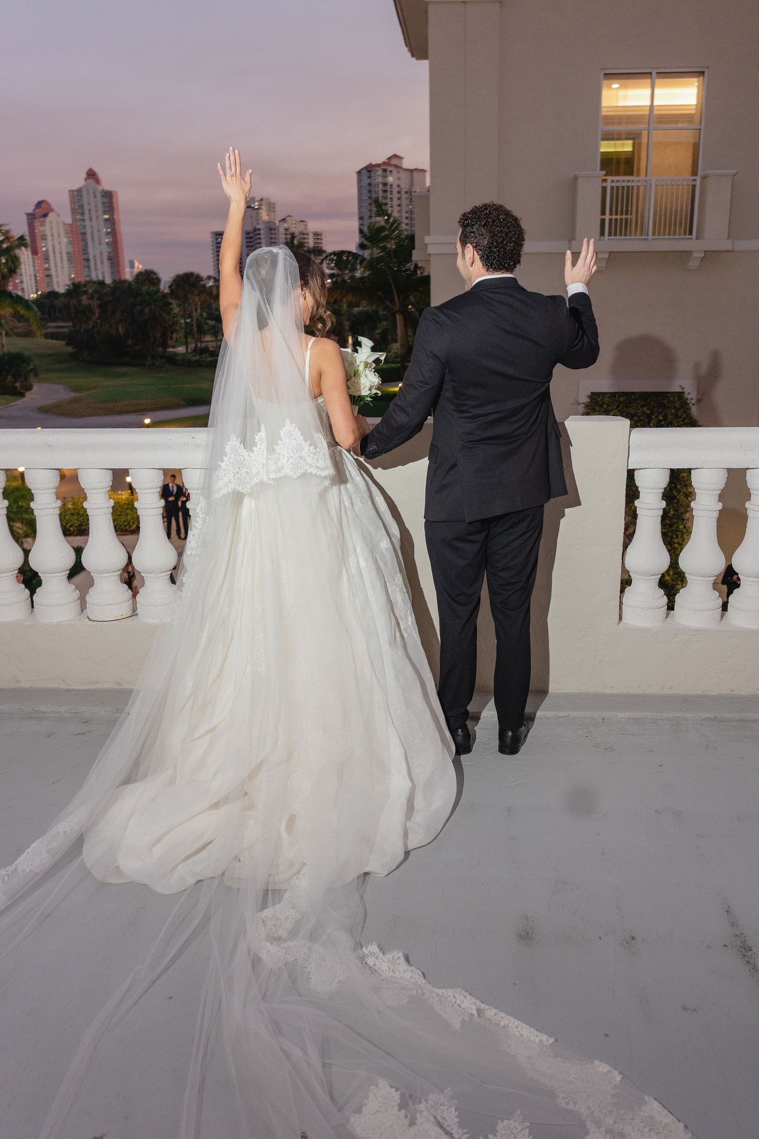 Bride and groom wave from a balcony at sunset, bride wearing a long white gown with flowing veil.
