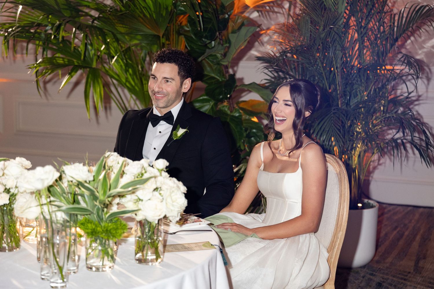 Bride and groom smiling at sweetheart table with white floral arrangements and tropical greenery at elegant wedding reception.
