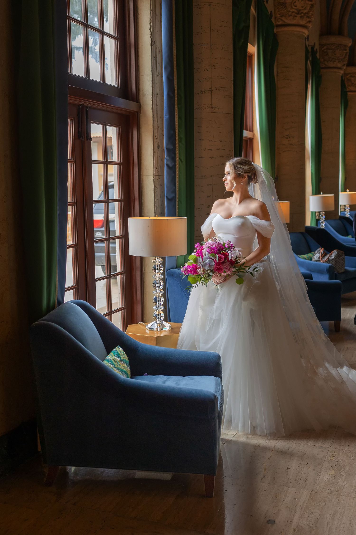 Elegant bride in white gown holding colorful bouquet stands by tall windows in a luxurious blue-furnished venue.