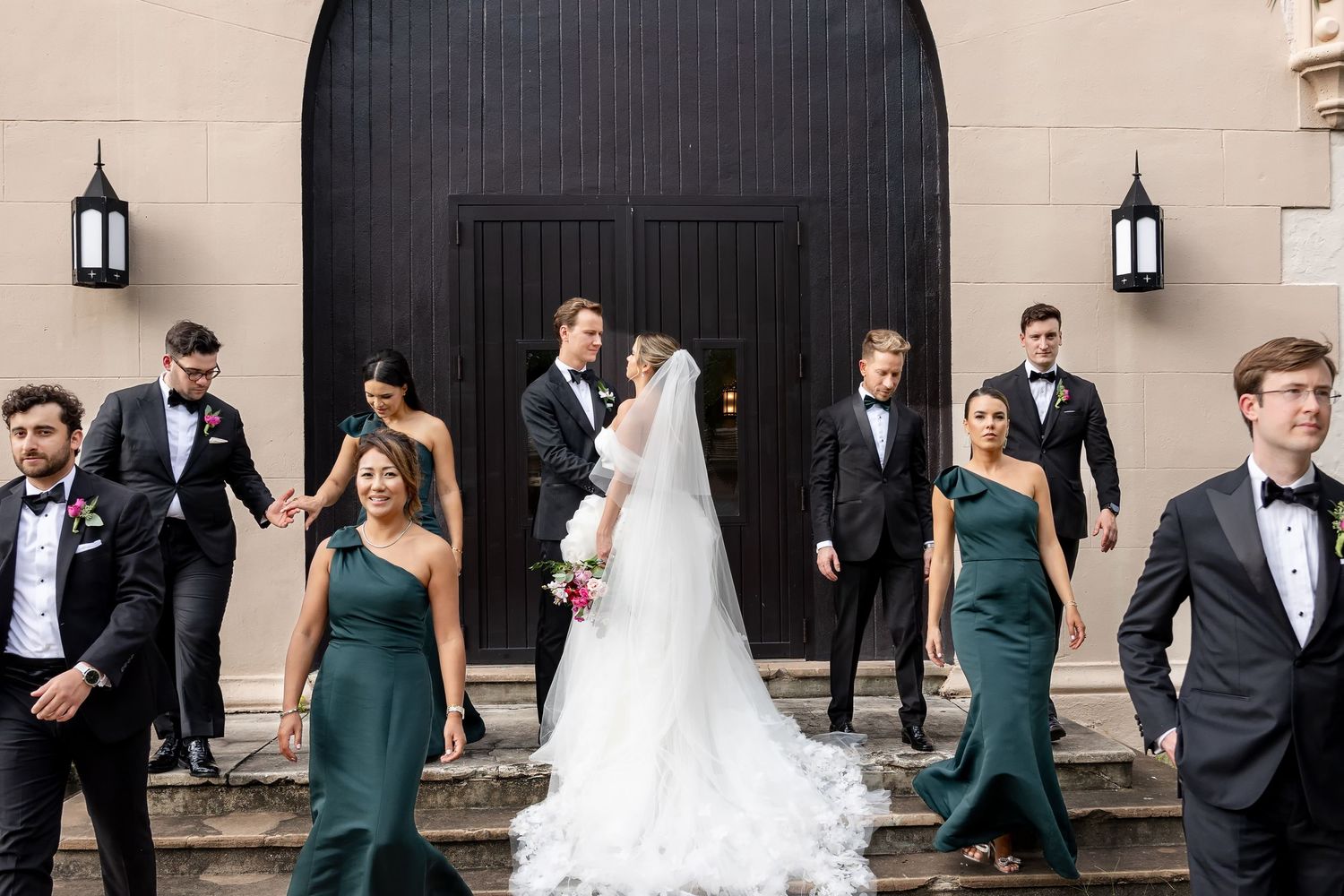 Wedding party in elegant attire, bridesmaids in emerald gowns and groomsmen in black tuxedos, outside a grand venue.