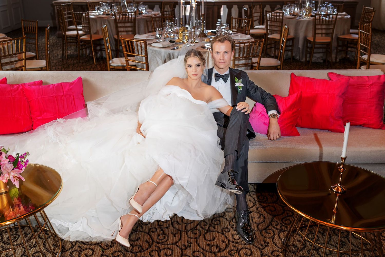 Bride in white gown and groom in gray suit pose together on a sofa at elegant wedding venue with red accents.