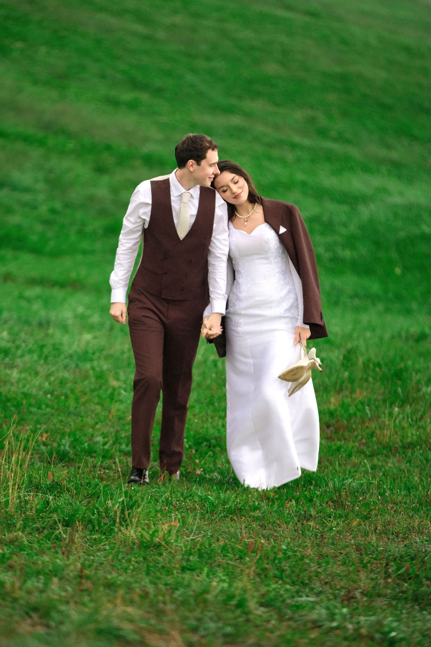 Newlywed couple walking hand-in-hand on green grass, bride in white gown with groom in brown suit.