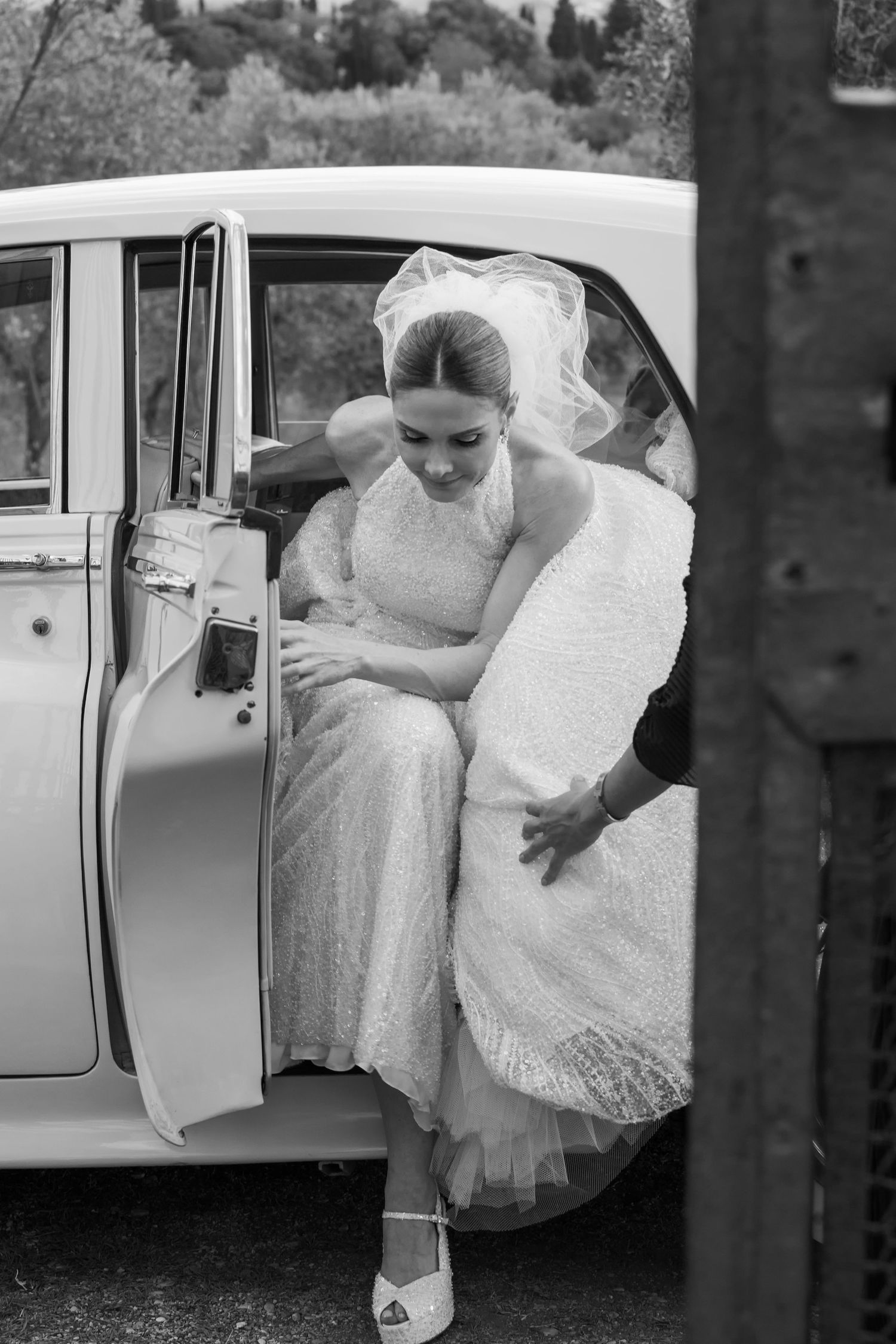 Classic black and white photo of bride stepping out of vintage car in lace wedding dress.