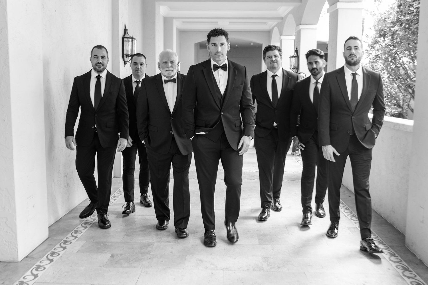 Group of seven men in black suits walking together in a elegant hallway, black and white wedding photo.