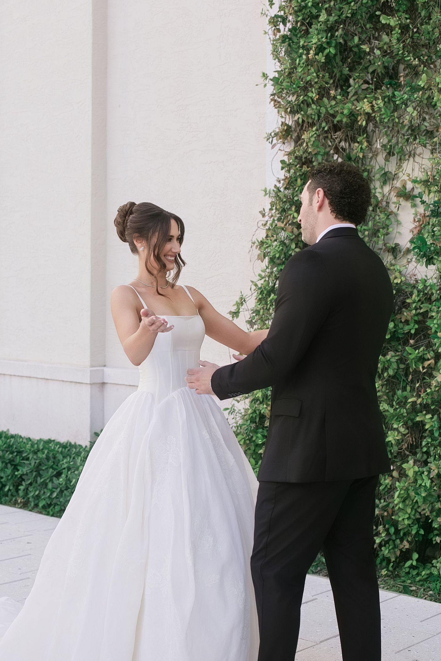 Bride in white ballgown and groom in black tuxedo share a tender moment outdoors beside ivy-covered wall.