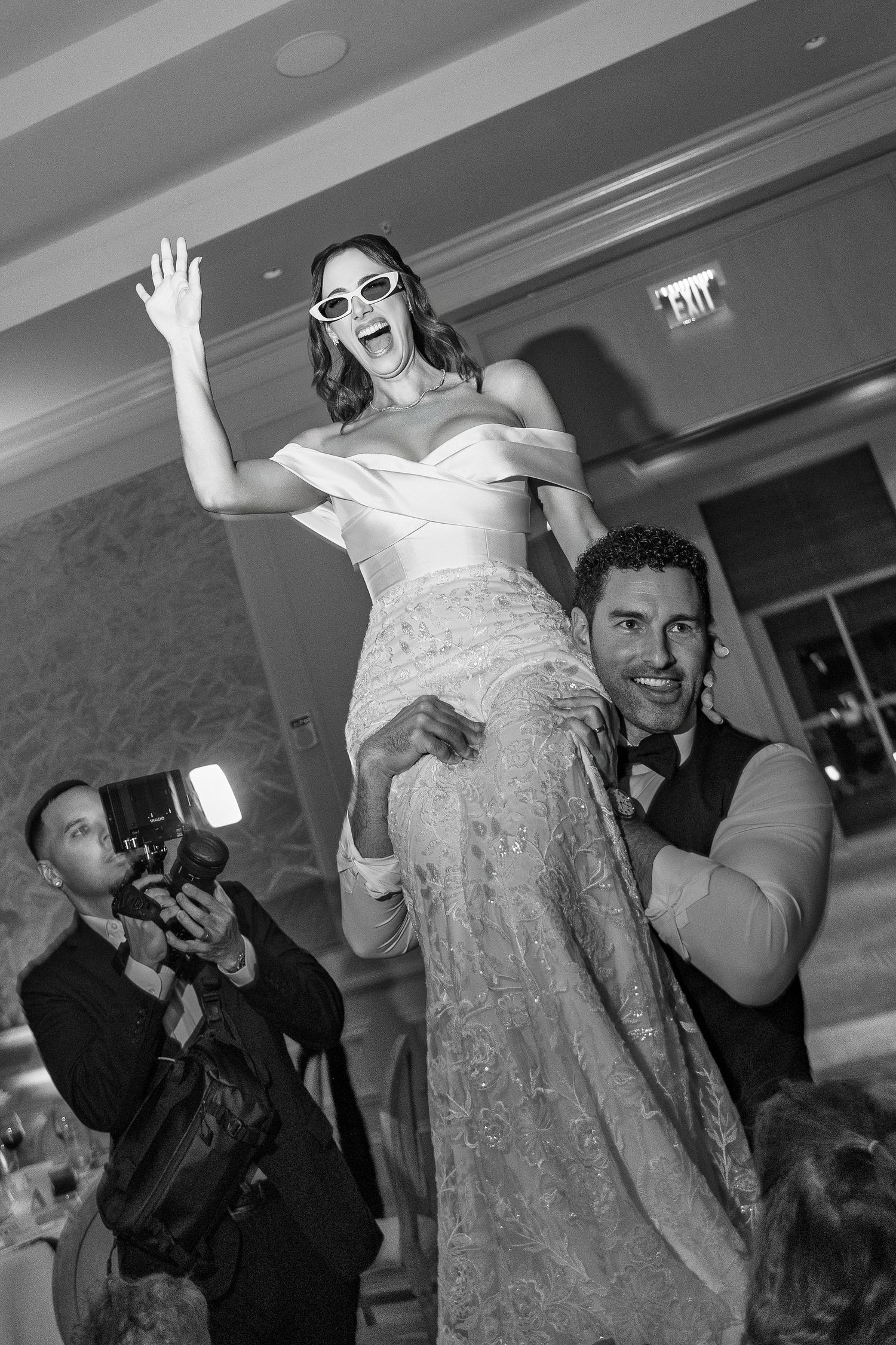 Bride in white gown and sunglasses lifted by groom at wedding reception, black and white photo.