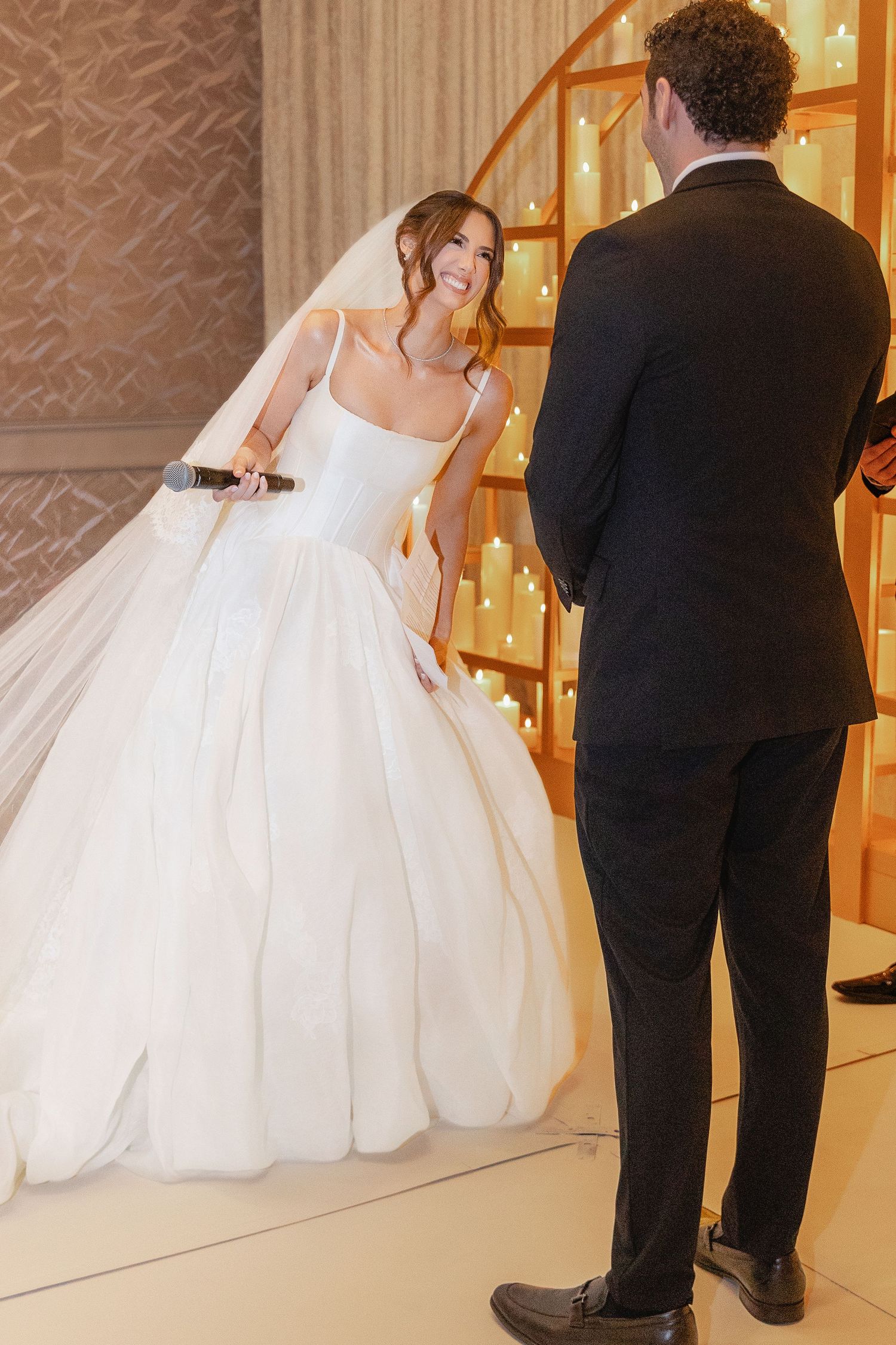 Bride in voluminous white ball gown smiles at groom near glowing staircase at wedding.