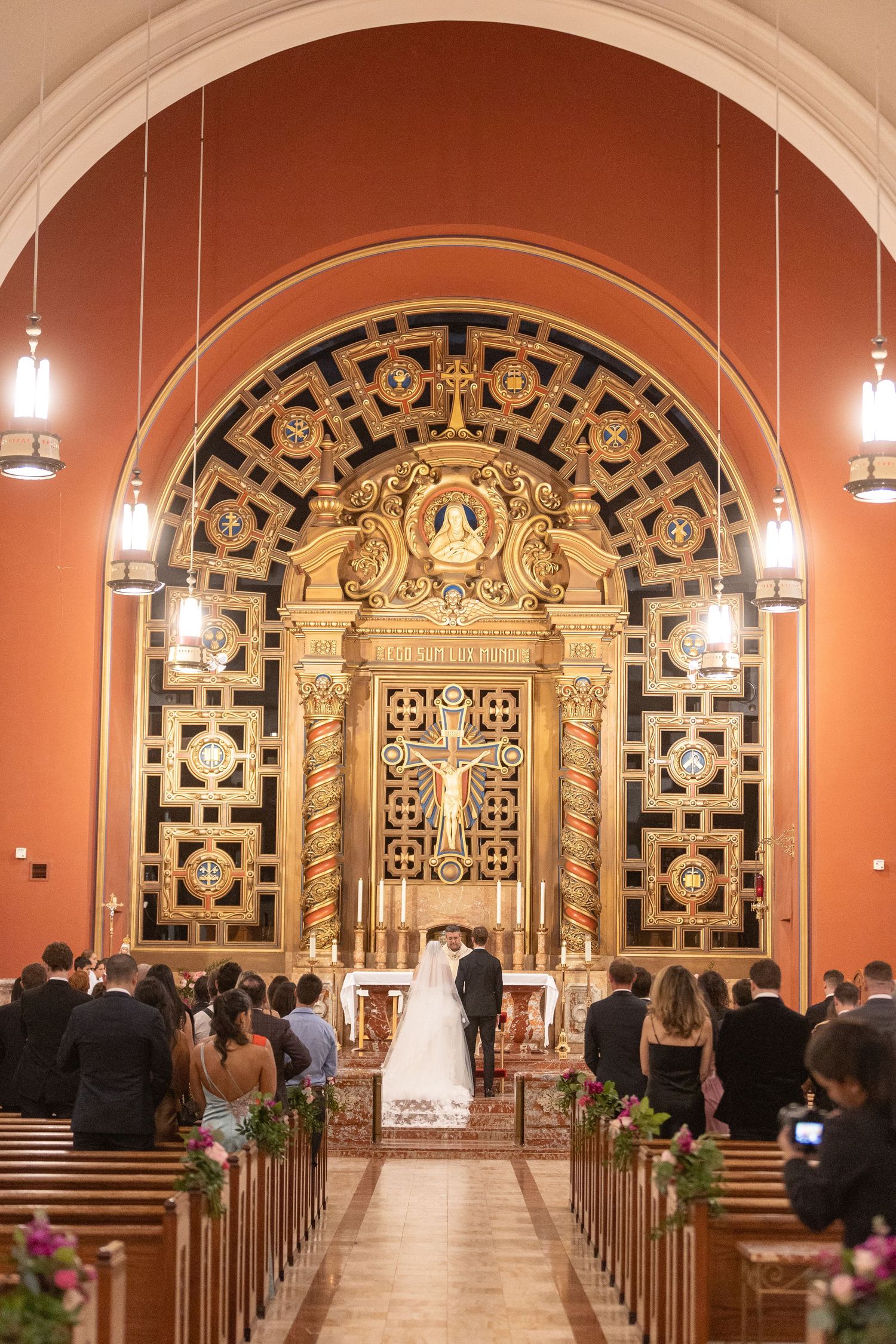 Bride and groom stand at ornate golden altar during wedding ceremony in elegant red Catholic church with pews full of guests.