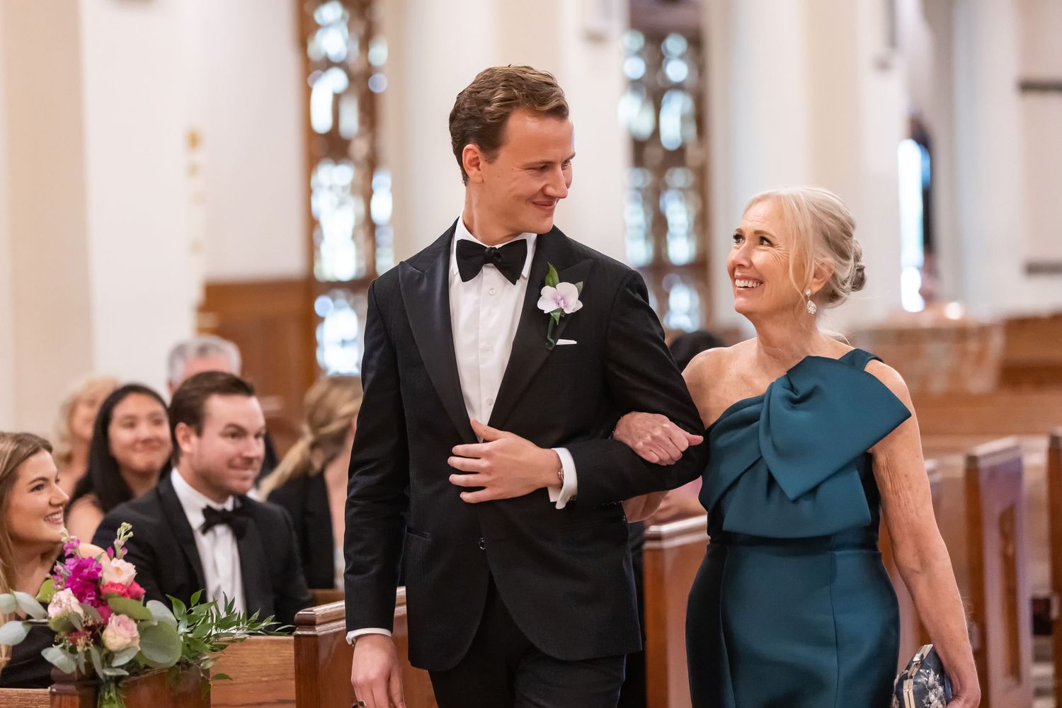 Groom in black tuxedo walks with elegant woman in teal gown down church aisle at wedding ceremony.