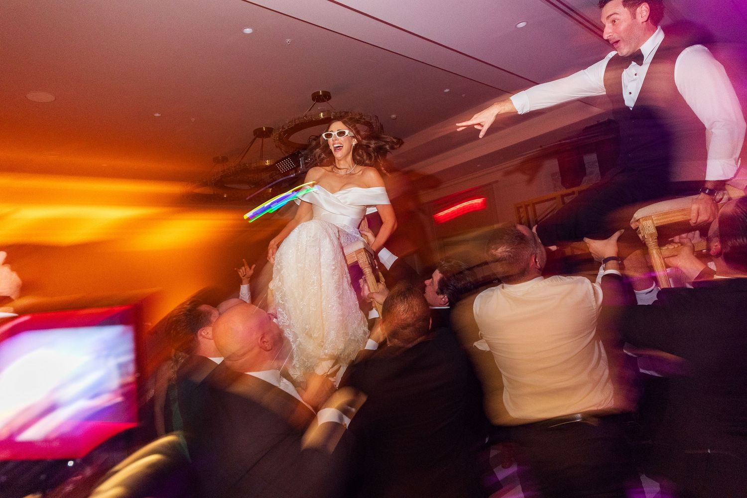 Bride in white gown lifted by guests on crowded wedding dance floor with warm ambient lighting.