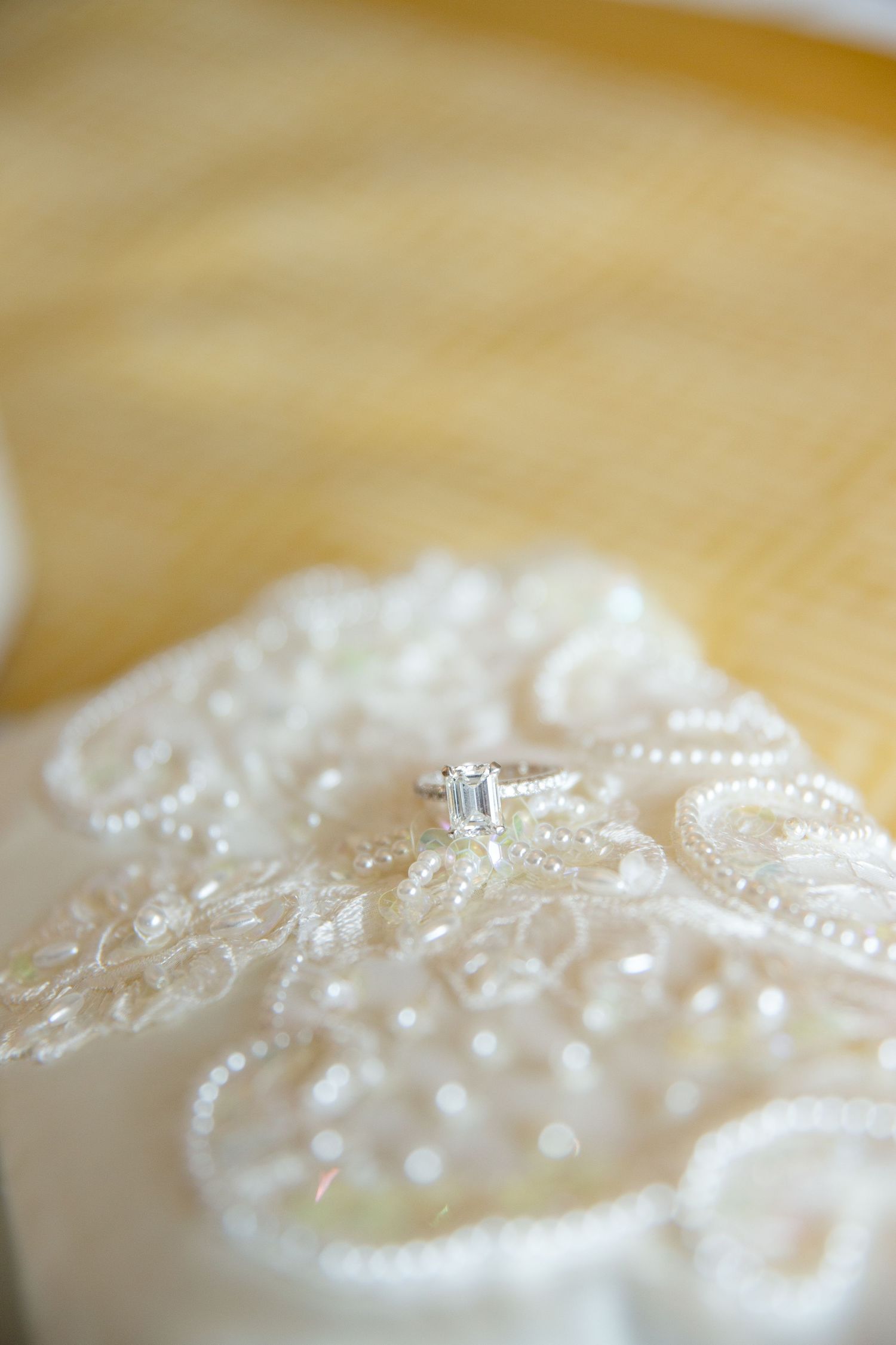 Elegant emerald cut diamond engagement ring resting on white beaded lace wedding dress fabric on wooden surface.