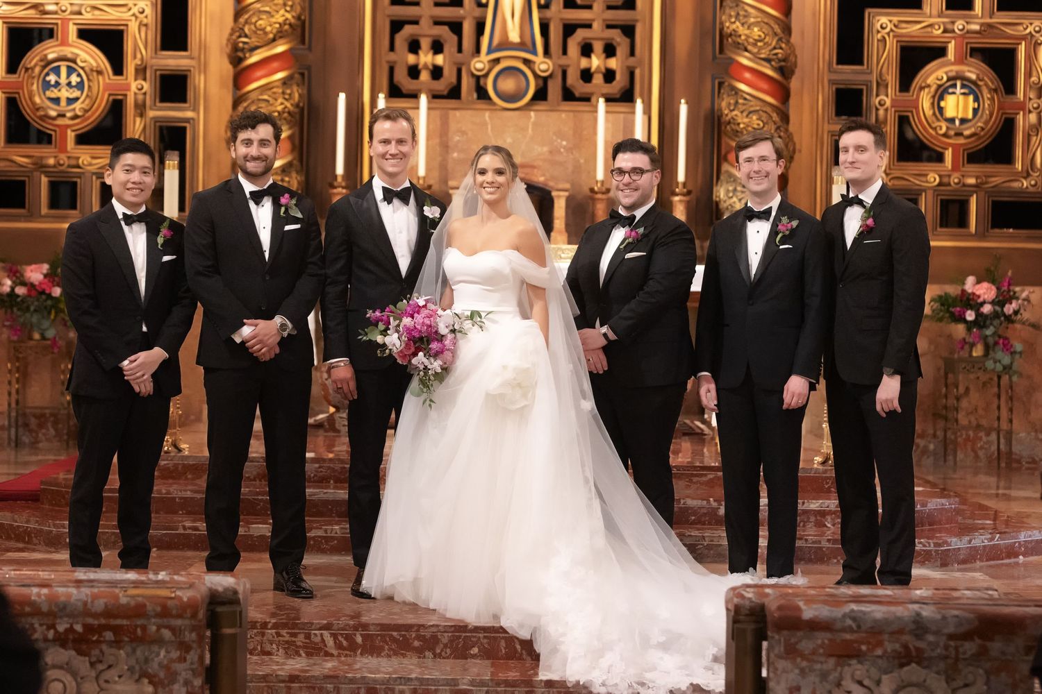Bride in white gown with groomsmen in tuxedos posing at ornate wedding ceremony altar with pink bouquet.