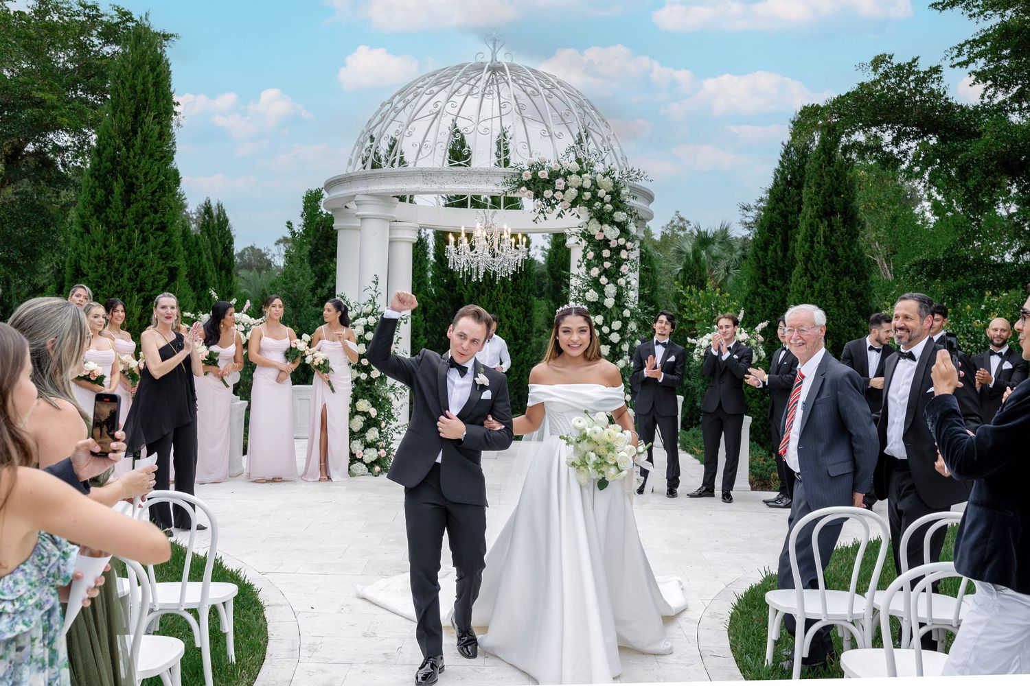 Outdoor wedding ceremony under white gazebo with floral arrangements and seated guests.