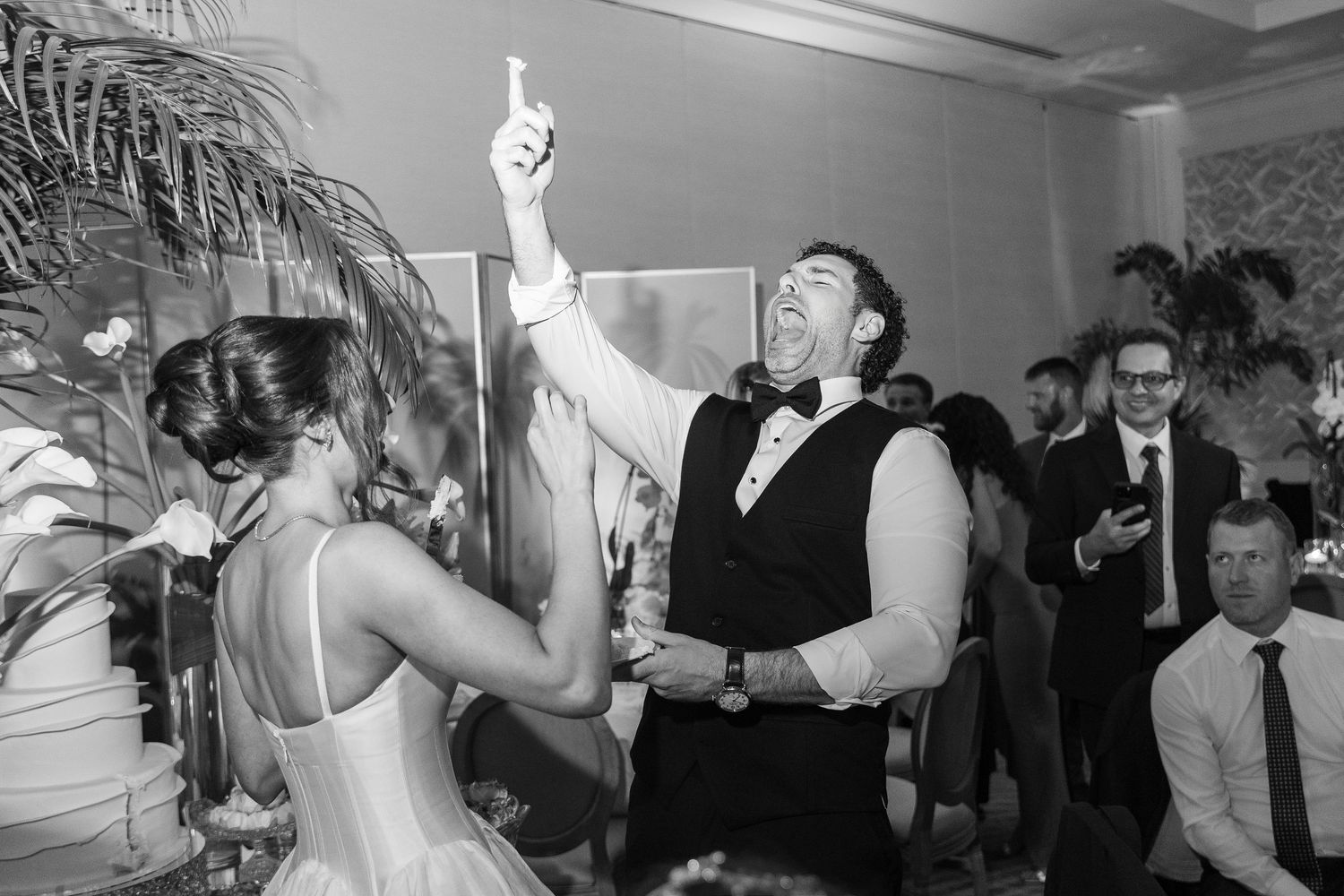 Black and white photo of a groom dancing joyfully with bride at wedding reception, guests clapping in background.