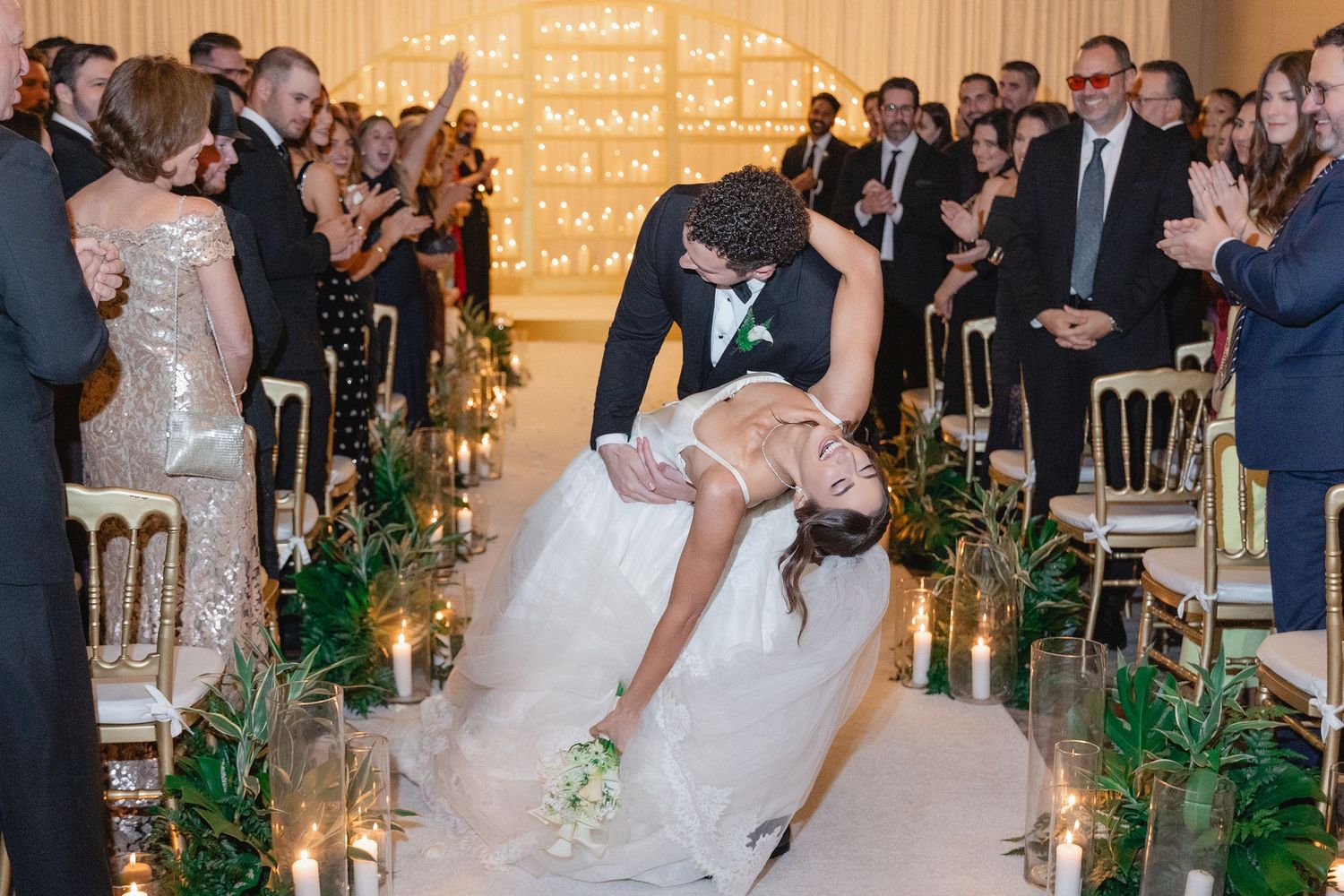 Groom dips bride in romantic wedding ceremony aisle kiss, surrounded by candles and cheering guests.