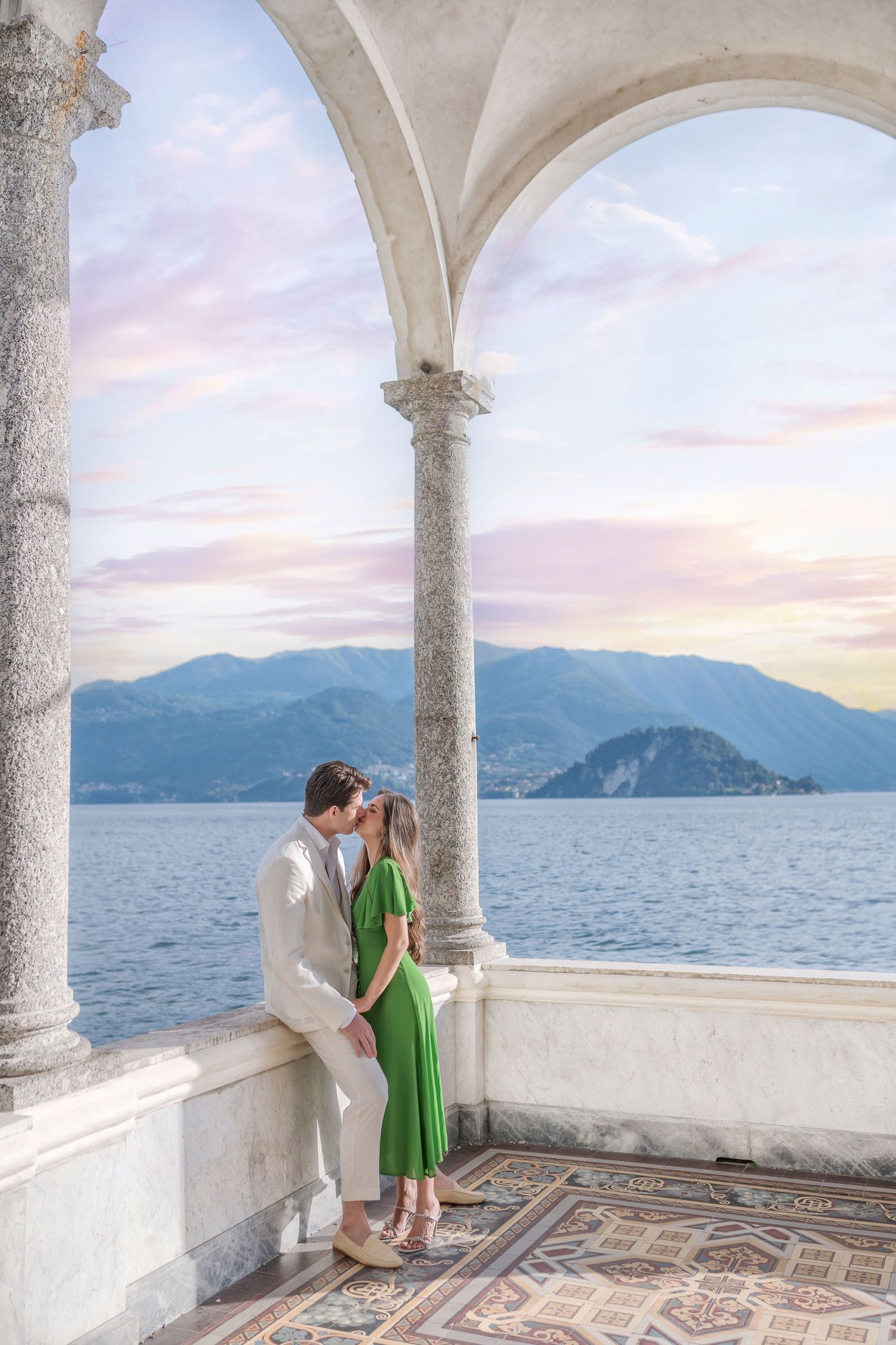 Romantic couple embraces on an elegant lakeside terrace with mountain views and classical stone columns.