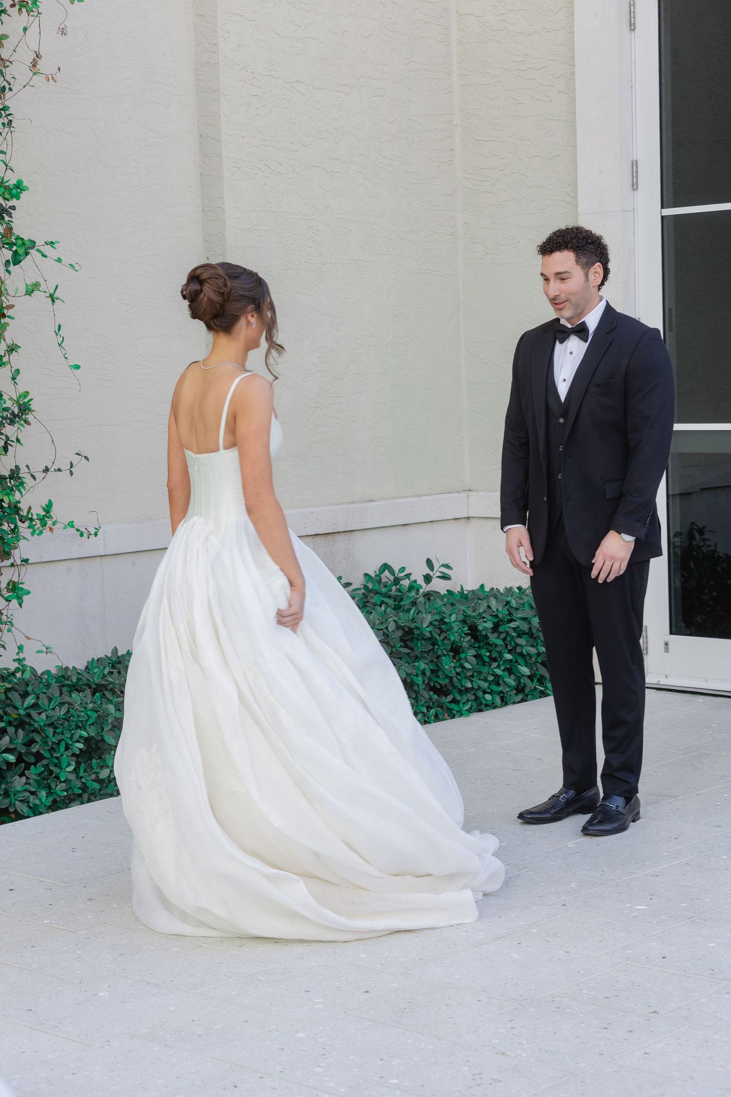 Bride in elegant white ball gown shares a first look moment with groom in black tuxedo outdoors.