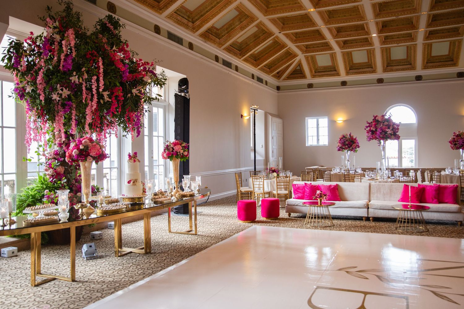 Elegant wedding reception hall with pink floral arrangements, gold accents, white dance floor, and fuchsia cushioned seating.