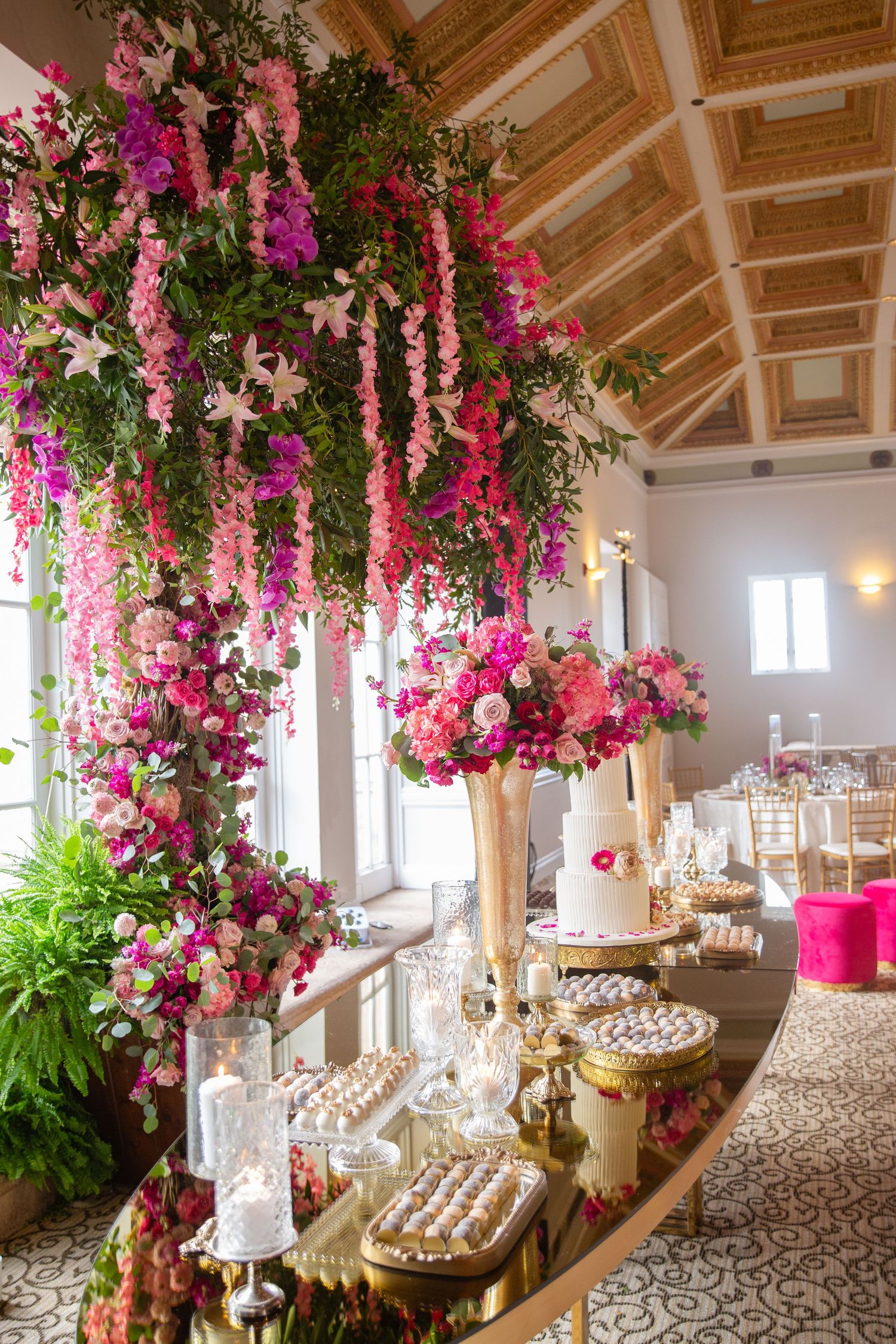 Elegant wedding dessert table adorned with cascading pink and fuchsia floral arrangements in a grand ballroom venue.