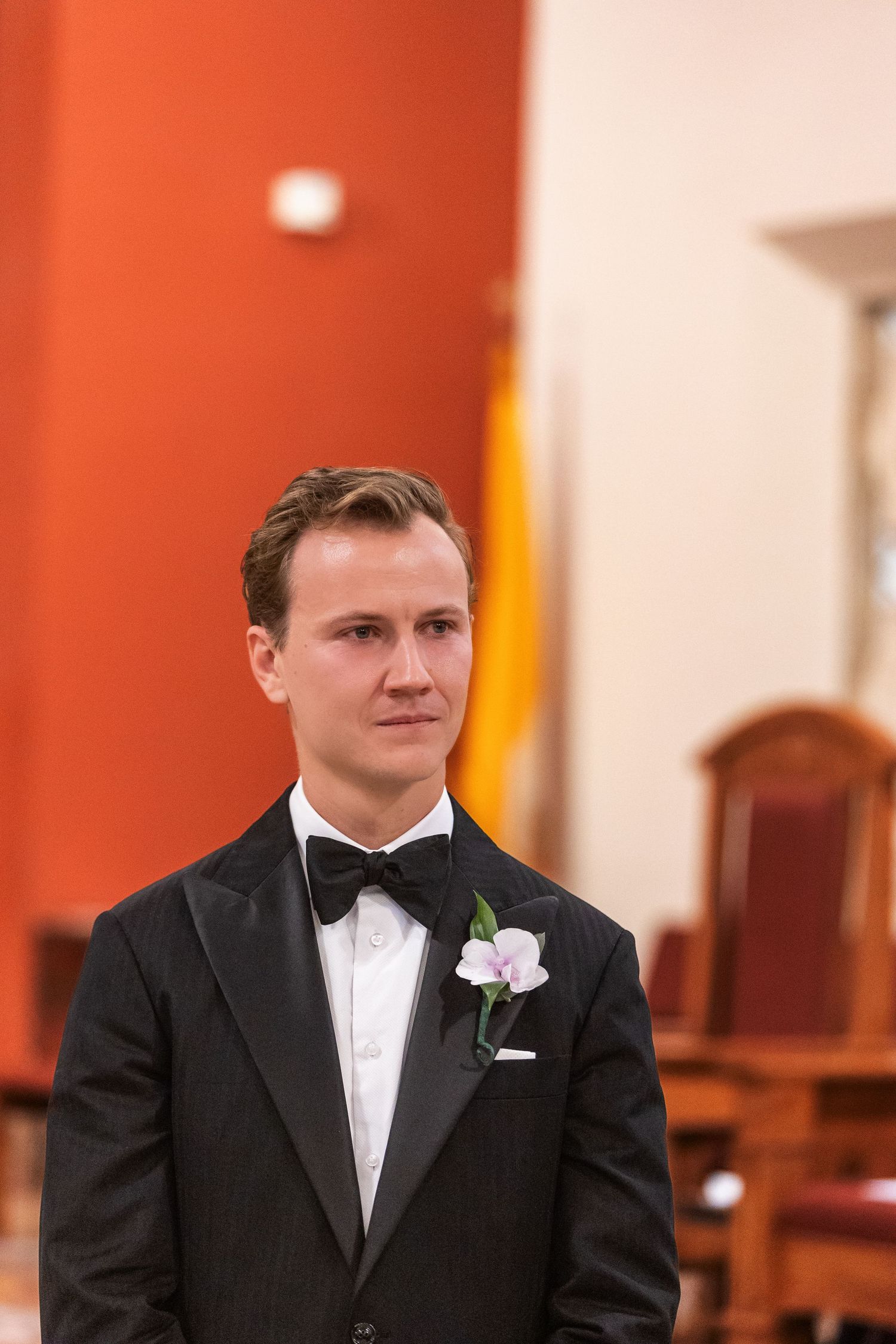 Groom in black tuxedo with bow tie and white orchid boutonniere standing in church during wedding ceremony.