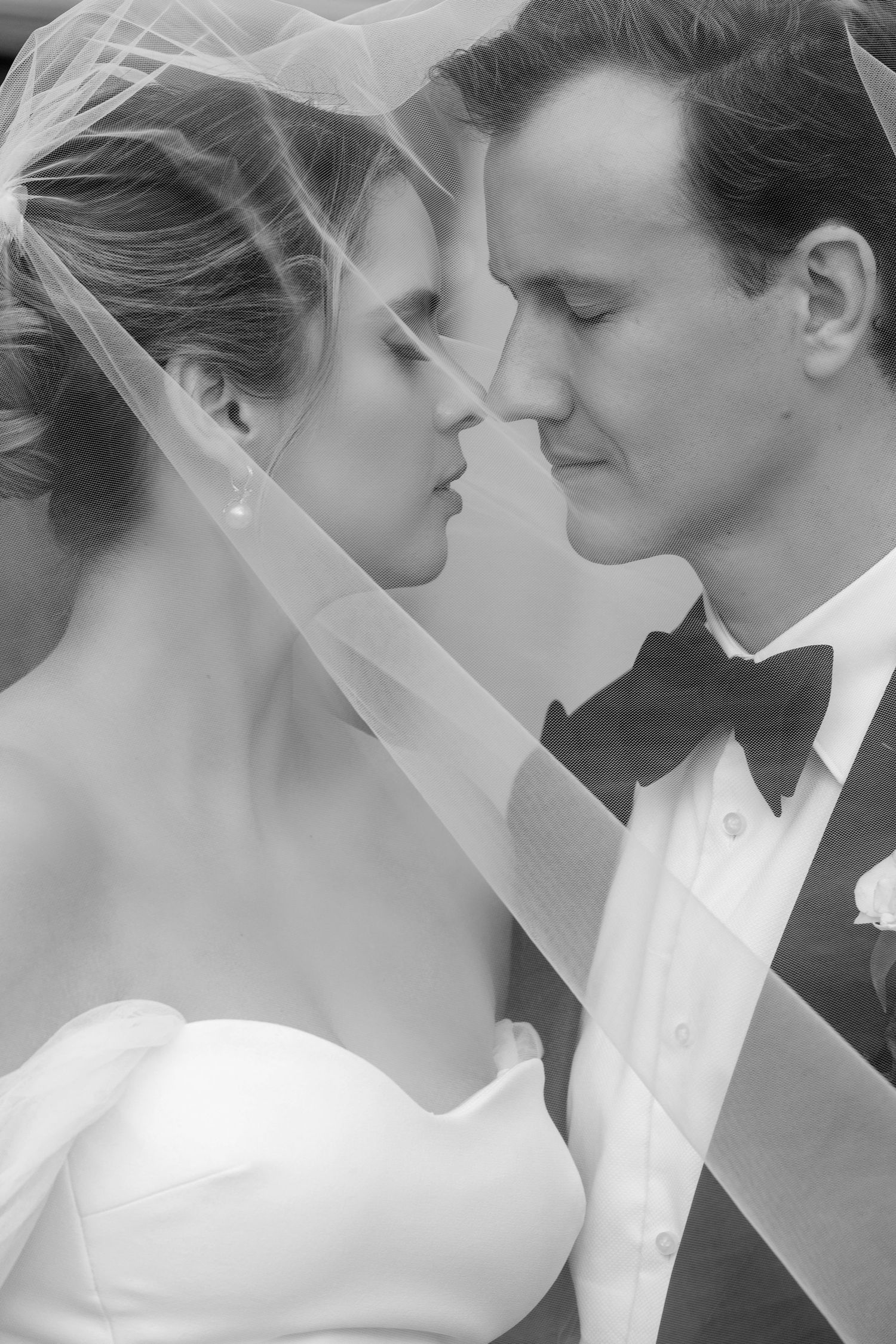 Black and white wedding photo of bride and groom sharing an intimate moment under a veil in formal attire.