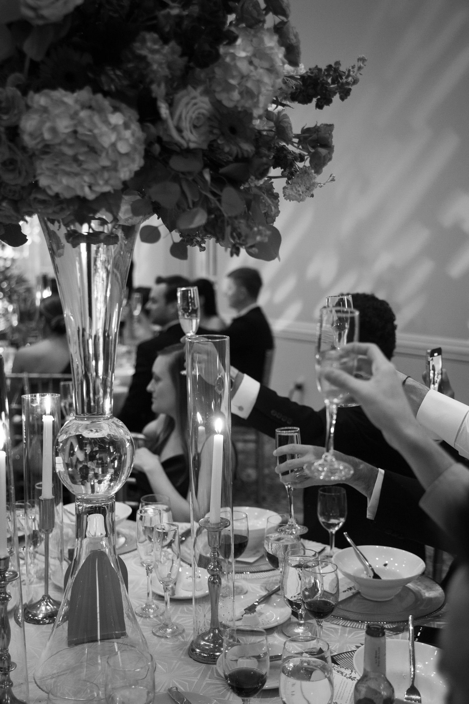 Black and white photo of elegant wedding reception table with tall floral centerpiece, candles, crystal glasses, and guests toasting.