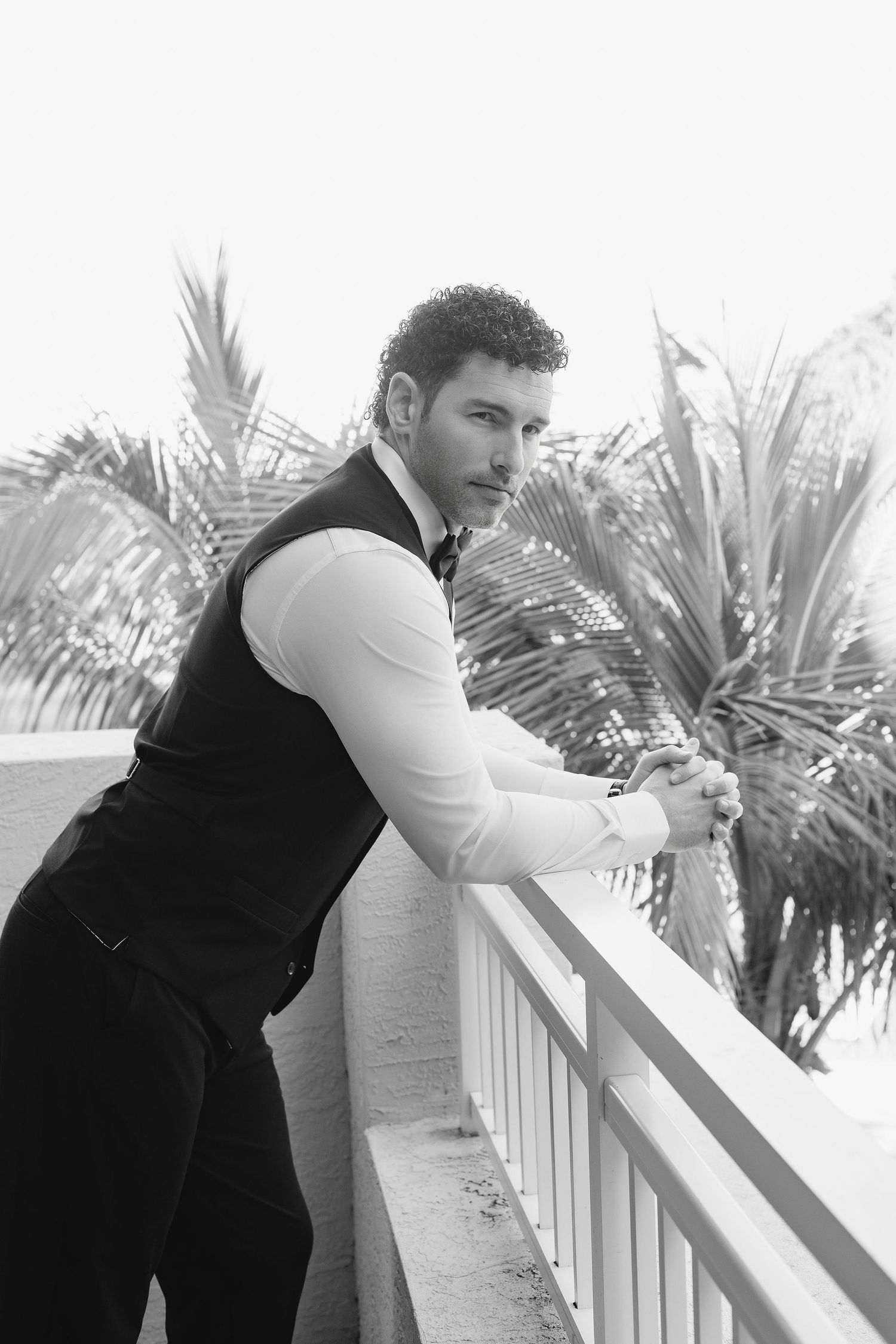A person in formal attire leans thoughtfully against a white railing with tropical palm trees in the background.