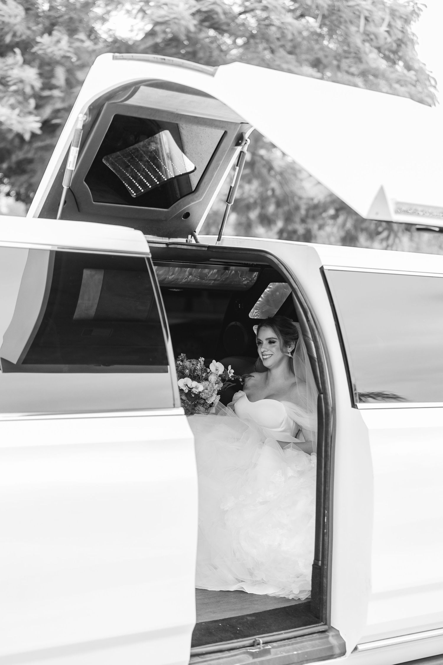 Black and white photo of a bride in a strapless gown holding flowers while seated inside a white stretch limousine.