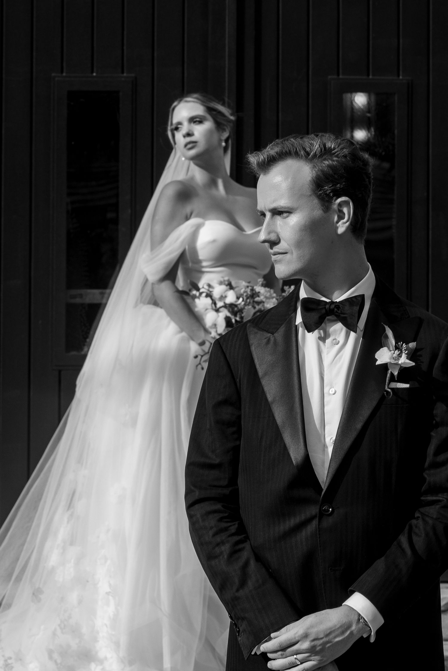 Black and white wedding photo of groom in tuxedo with bow tie, bride in flowing gown and veil standing behind him.