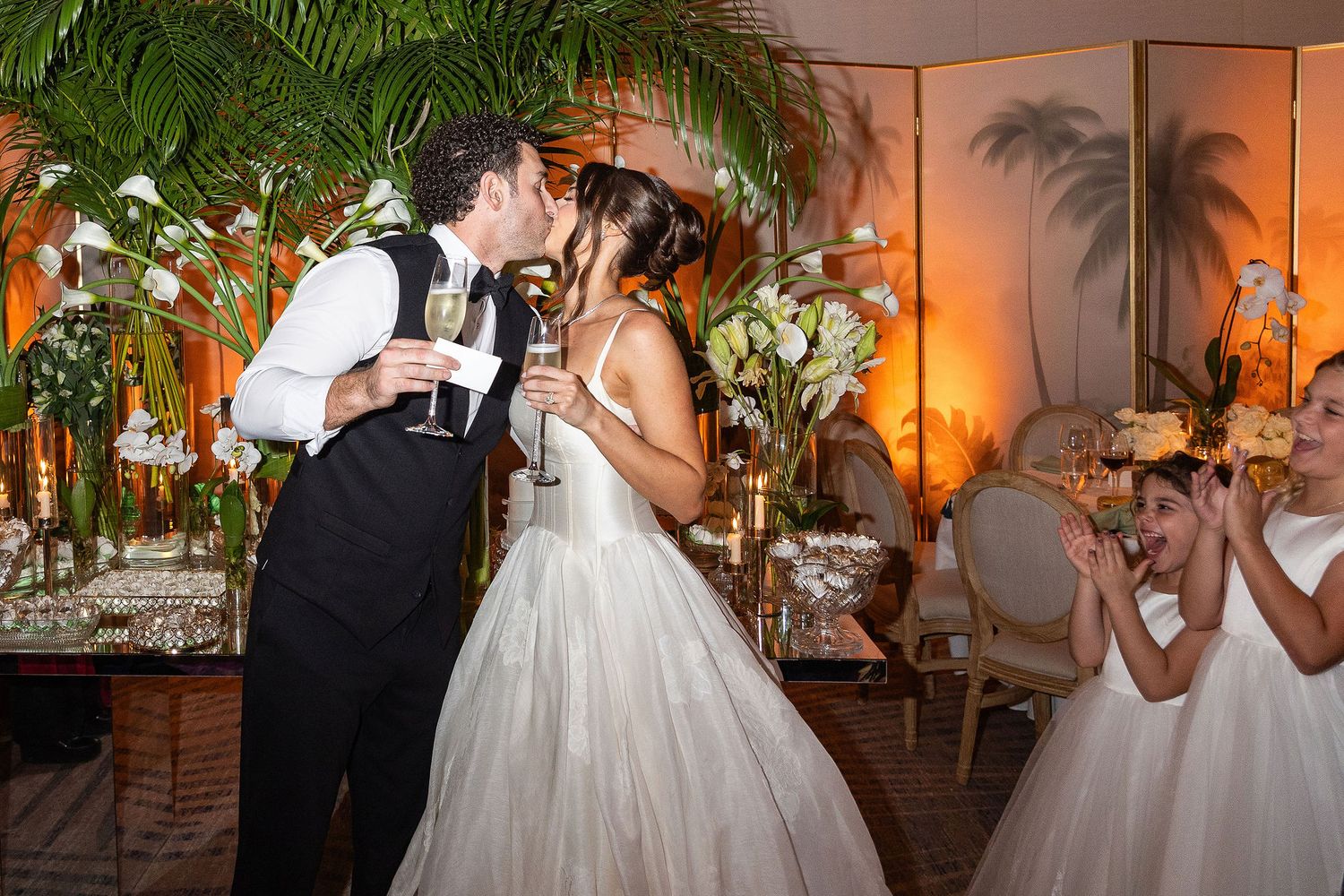 A couple shares their first dance at an elegant evening wedding reception with palm trees and warm lighting.