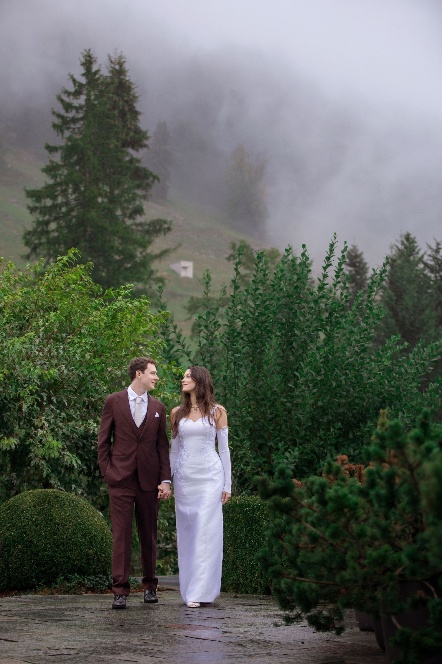 Bride and groom walk hand-in-hand through a misty mountain garden, she in white gown, he in burgundy suit.