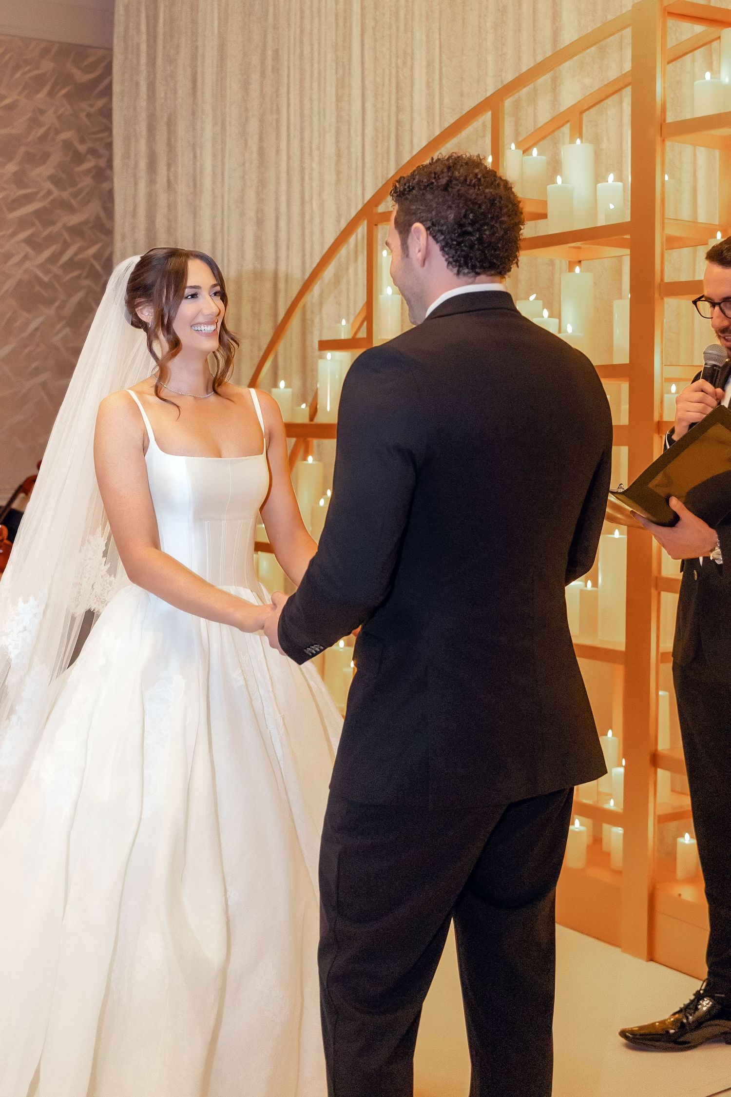 Bride in white ball gown and veil holds hands with groom in black tuxedo during indoor wedding ceremony.