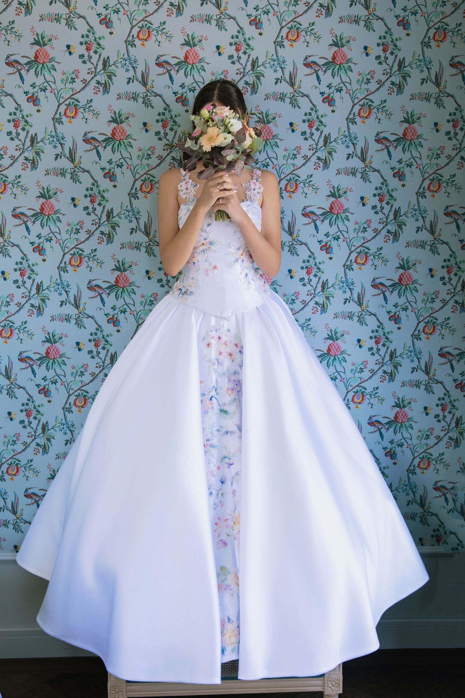 Bride in white ballgown with floral details holds bouquet covering face, standing before blue botanical wallpaper.