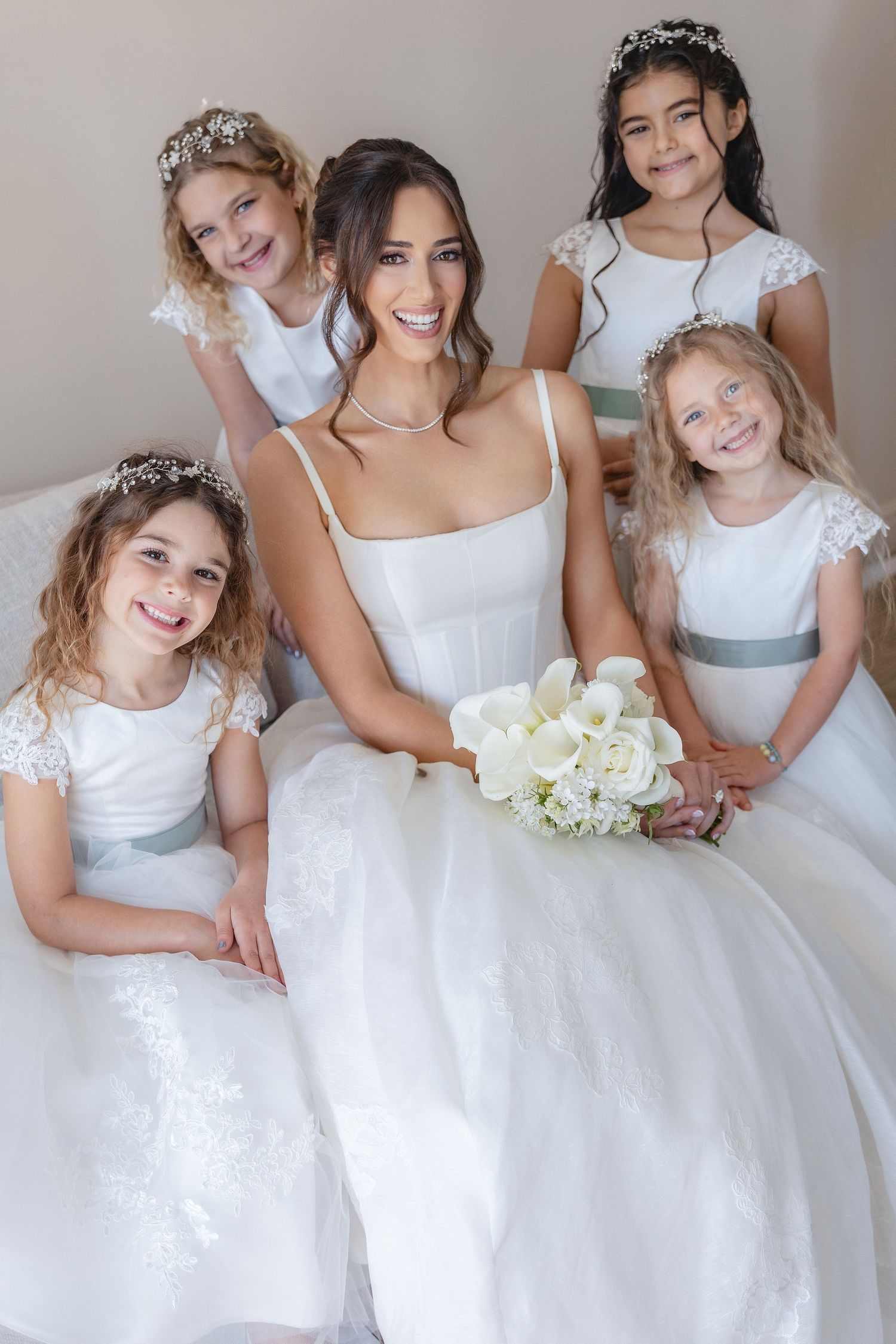 Bride in white gown holding bouquet poses with four flower girls in white dresses and tiaras at a wedding.