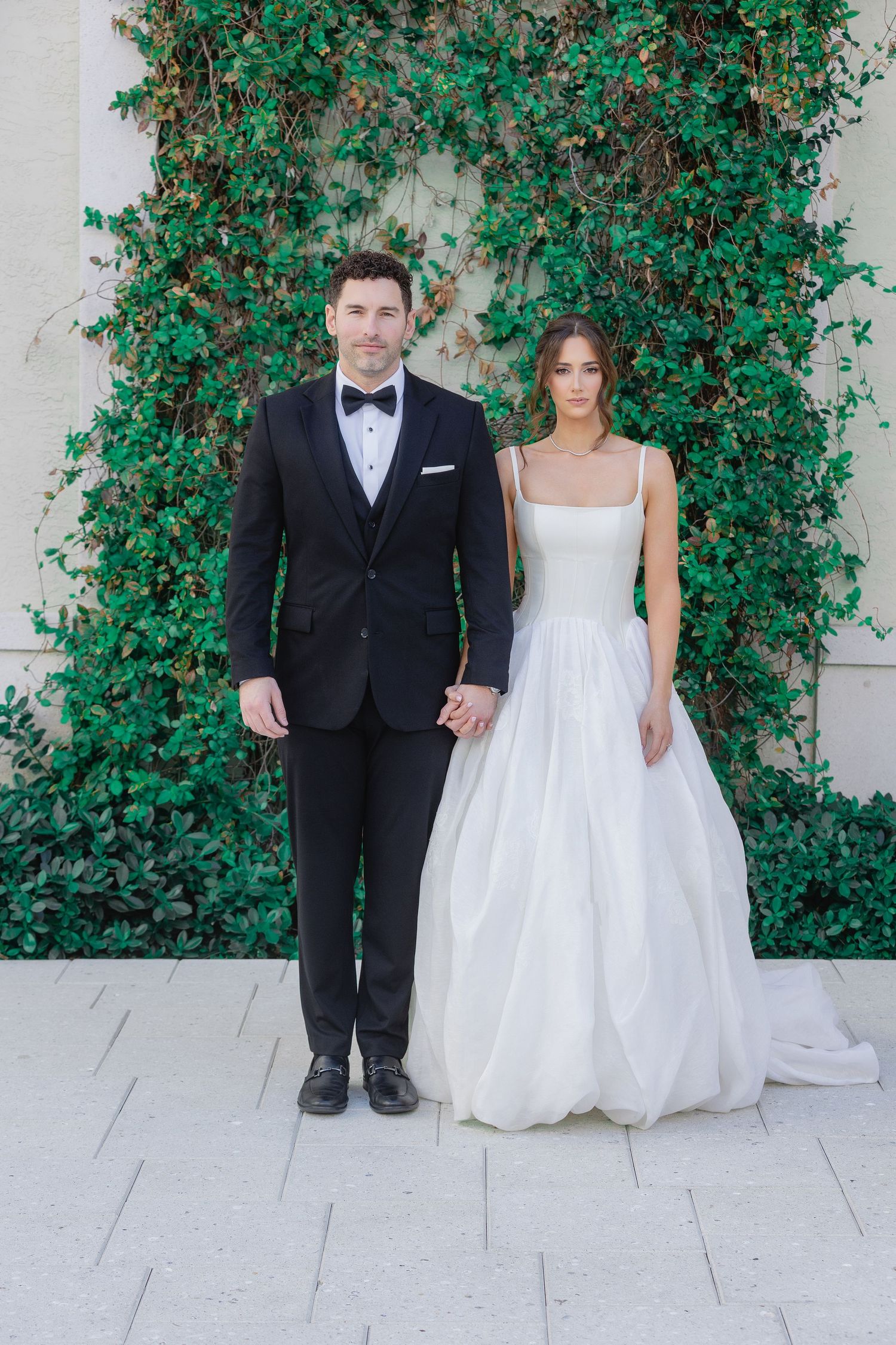 Bride in white ballgown and groom in black tuxedo hold hands before lush green ivy wall backdrop.