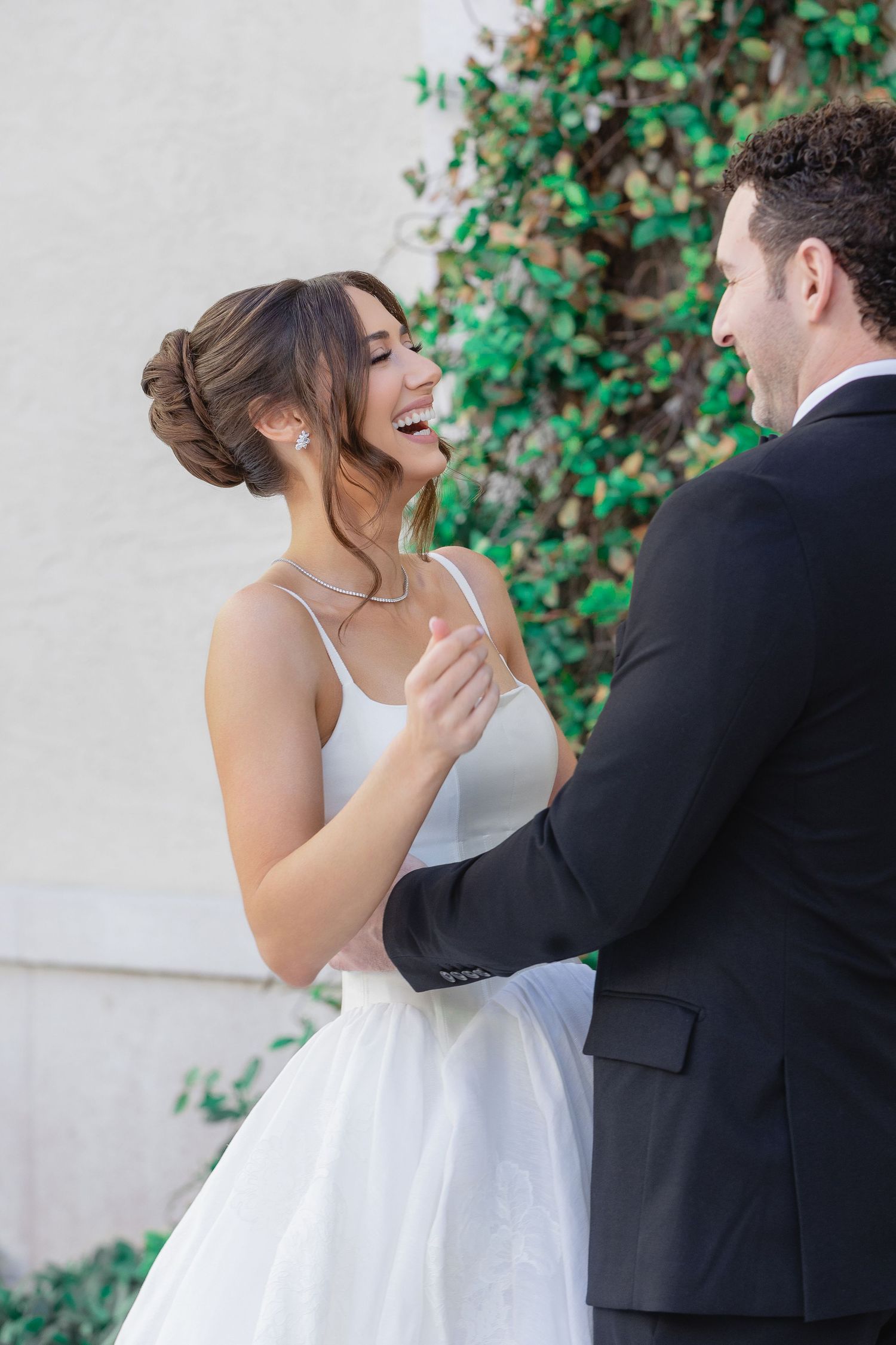 Bride in white gown laughs joyfully with groom in black suit during first look, ivy wall backdrop.