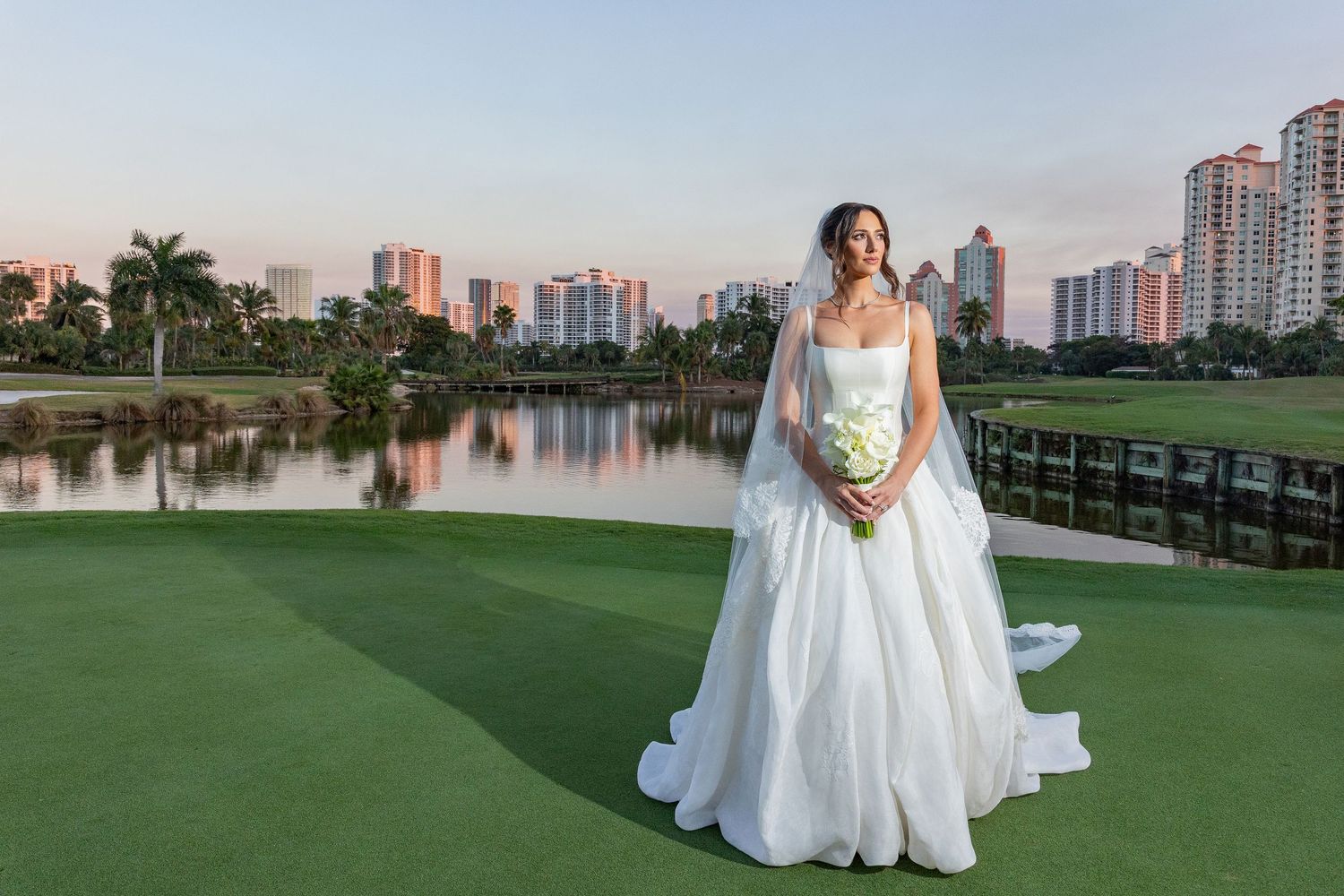 Elegant bride in white ball gown with veil holding bouquet on golf course with city skyline at sunset.
