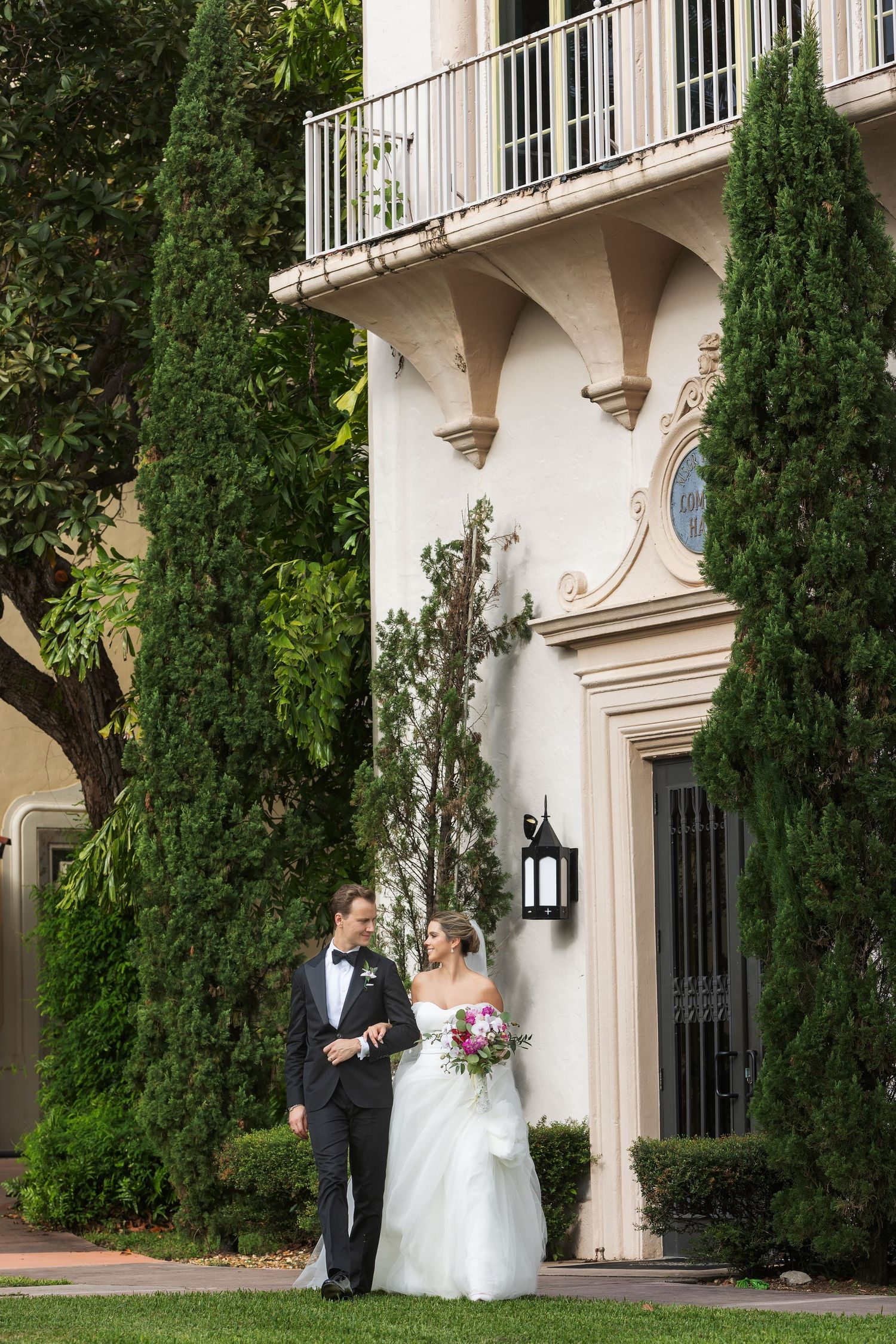 Bride and groom pose together in wedding attire outside an elegant white building with tall cypress trees and a balcony.
