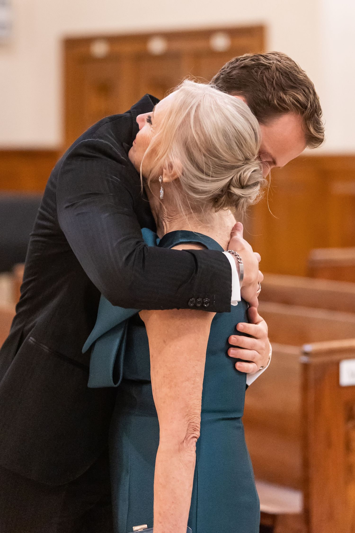 A young man in a dark suit embraces an older woman in a teal dress inside a church with wooden pews.