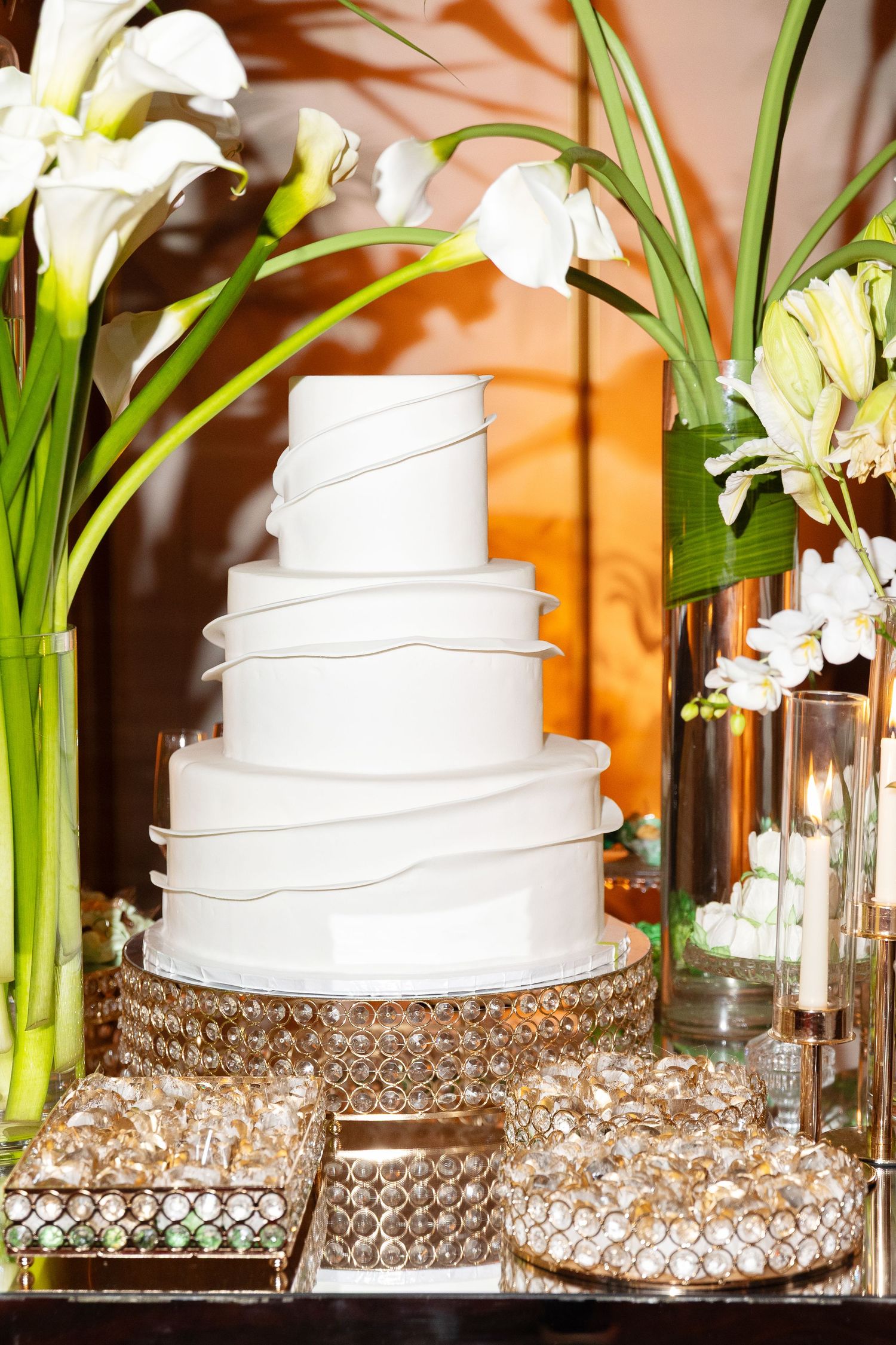 Elegant white four-tier wedding cake with ribbon details on crystal stand, surrounded by calla lilies and candles.