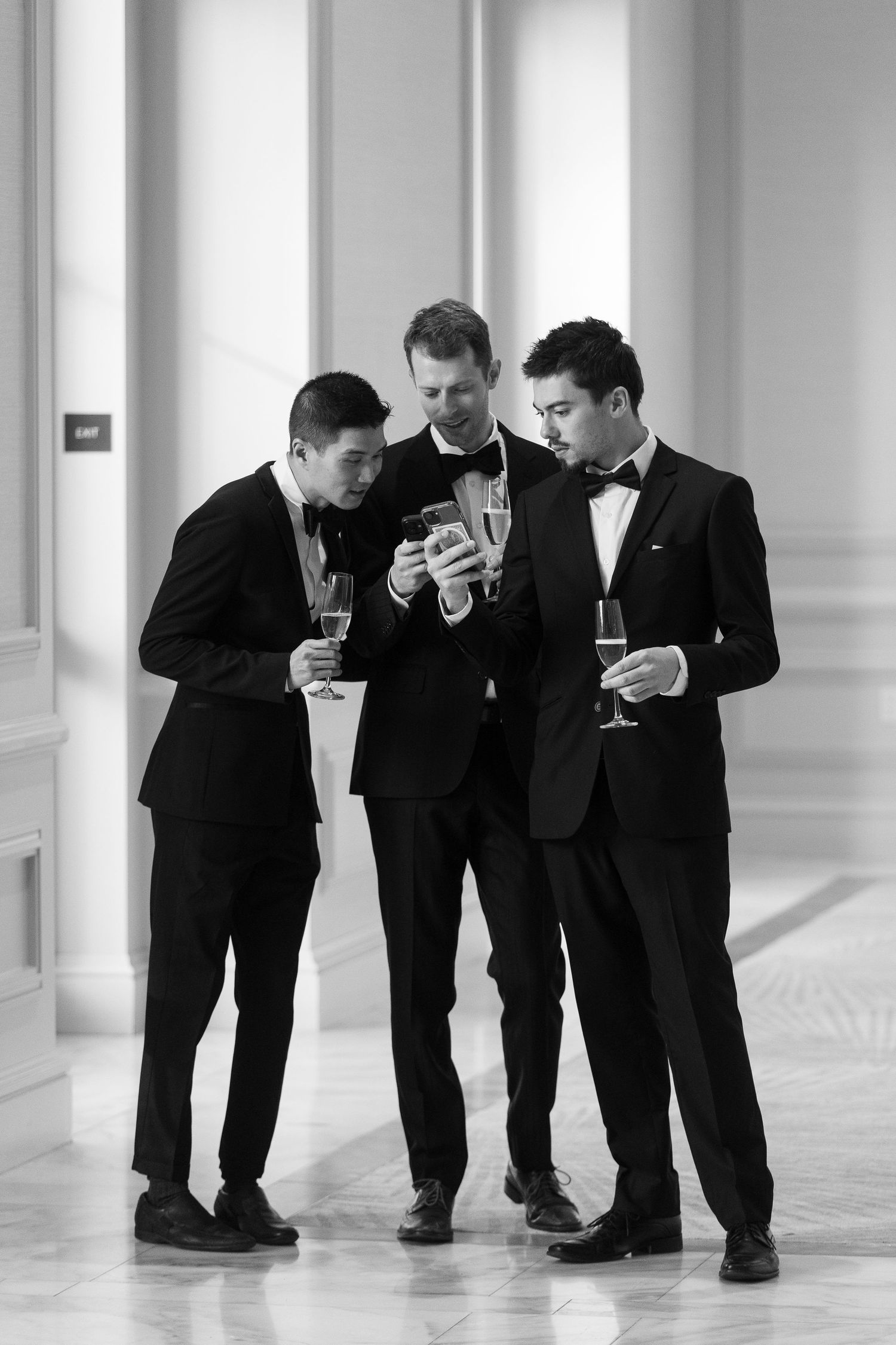Three men in tuxedos with bow ties holding champagne glasses while looking at a smartphone in a elegant hallway.