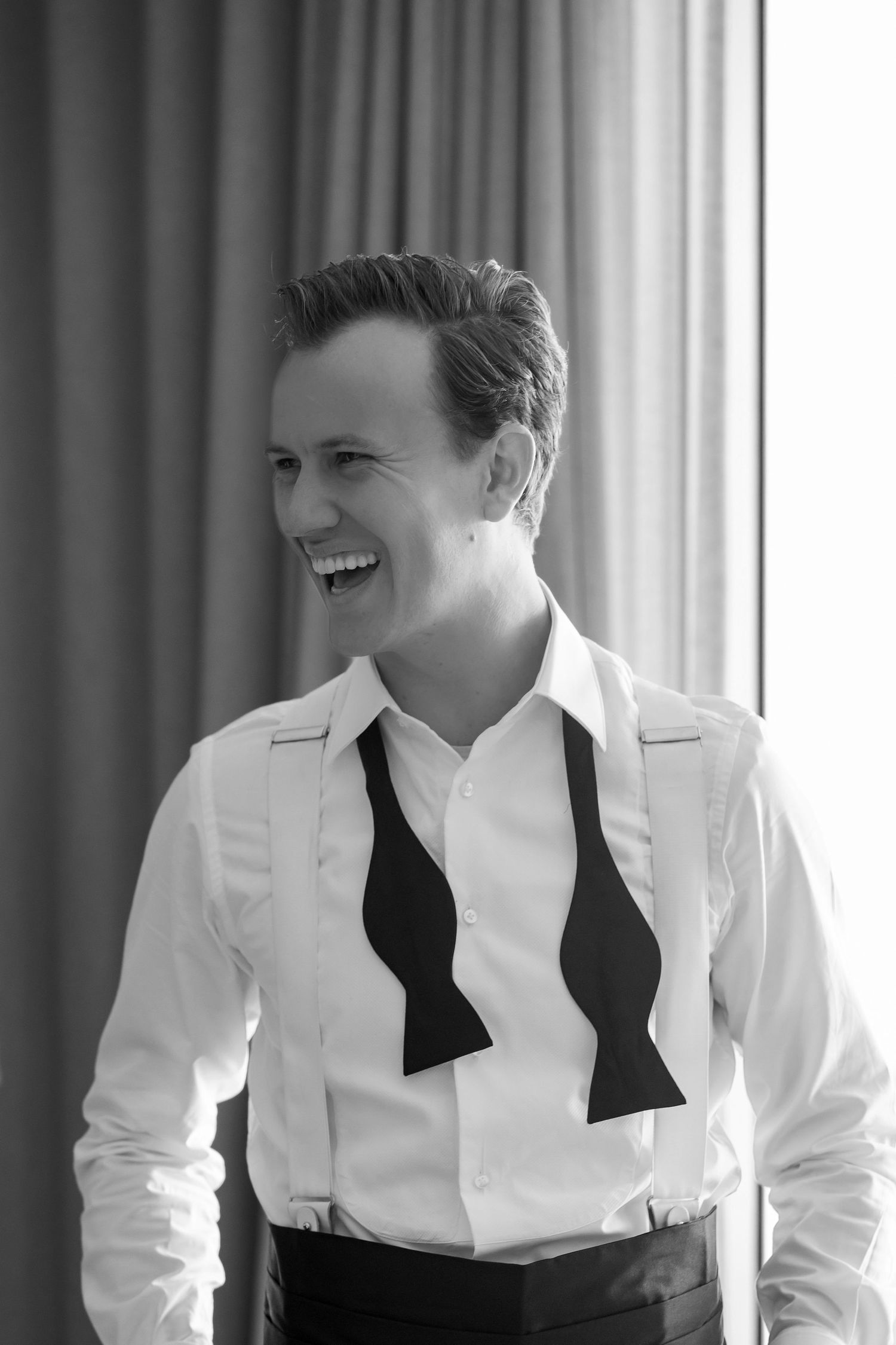 Black and white photo of a smiling person in a white shirt and bow tie, standing near a curtained window.