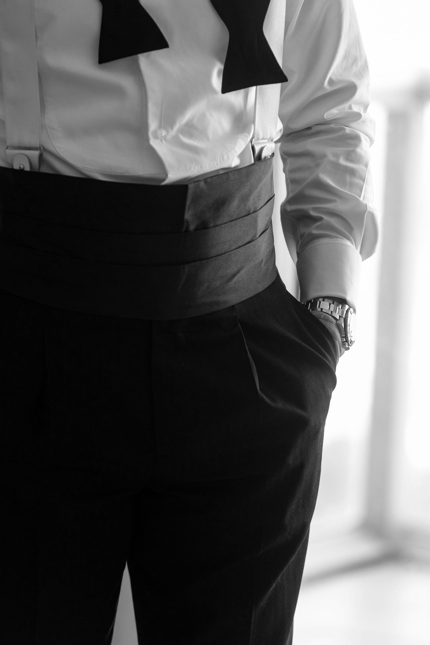 Black and white photo of a man in a tuxedo with bow tie, cummerbund, and dress watch, hands in pockets.