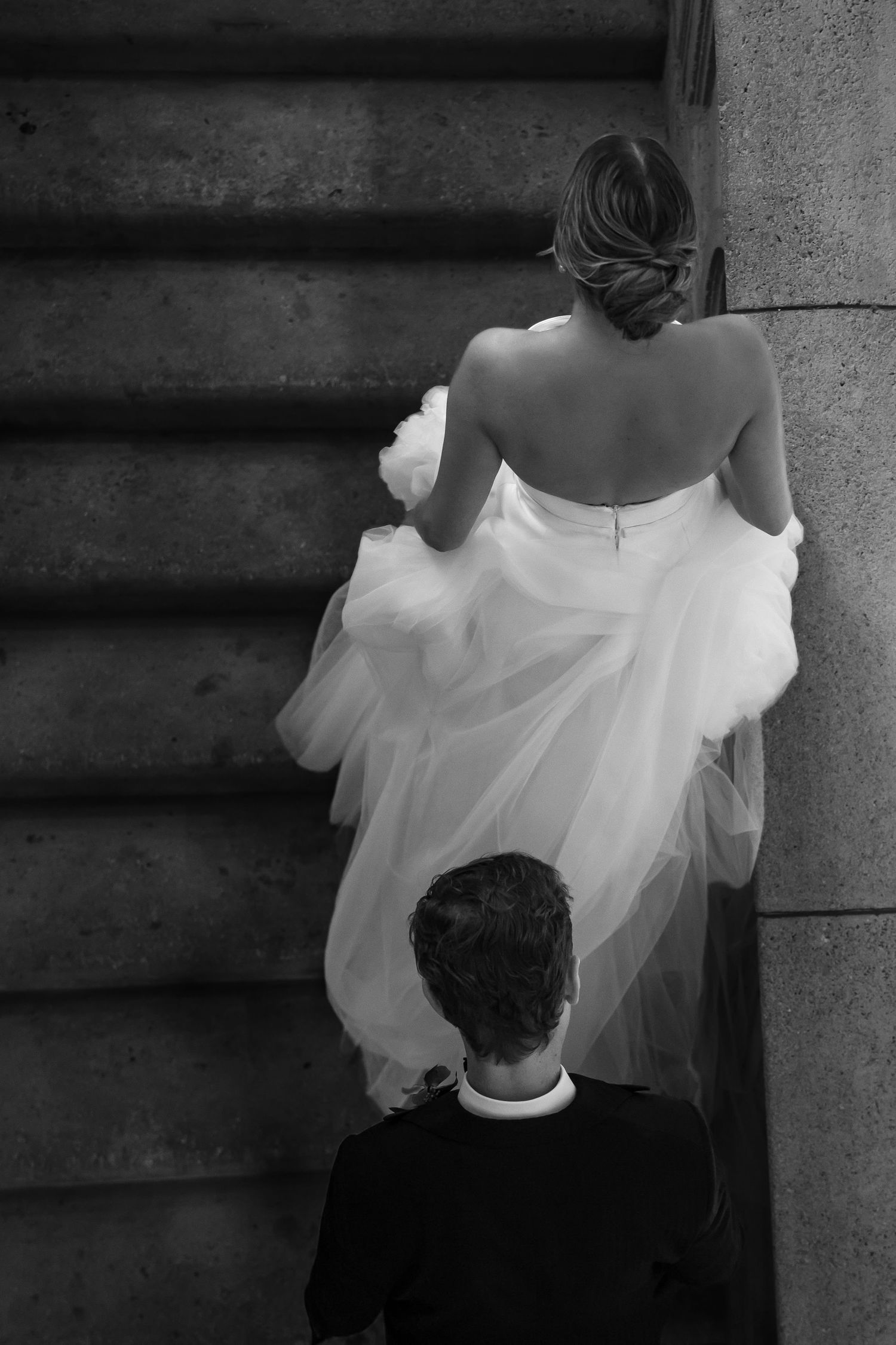Black and white photo of a bride in a strapless gown ascending stone stairs, followed by a groom in a dark suit.