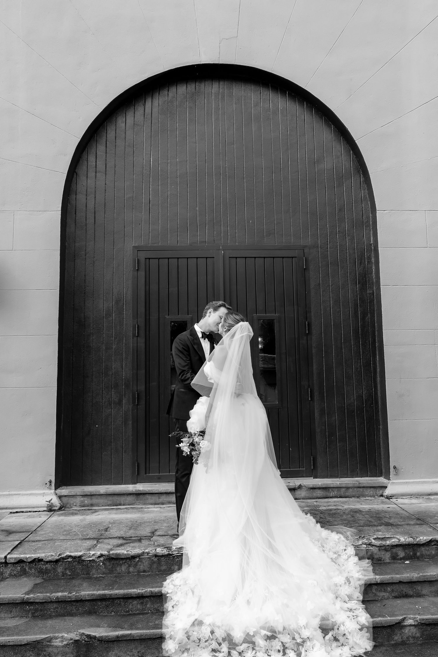Black and white photo of bride and groom kissing before large arched wooden doors, bride's cathedral veil trailing on steps.