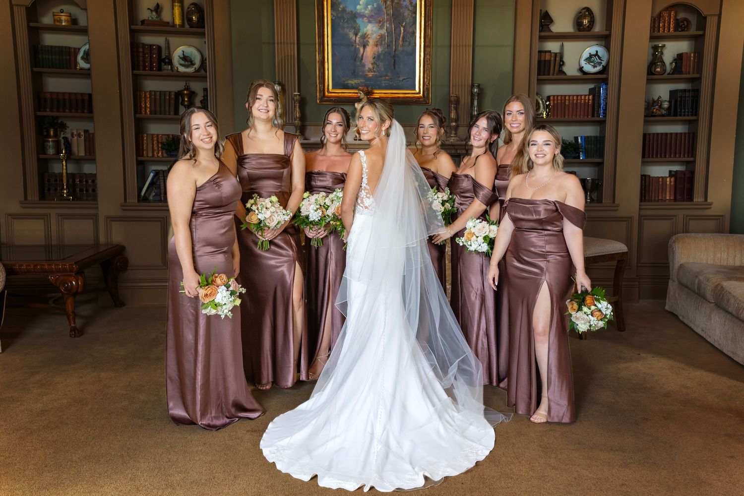 Wedding party in purple bridesmaid dresses poses with bride in elegant white gown at indoor venue.