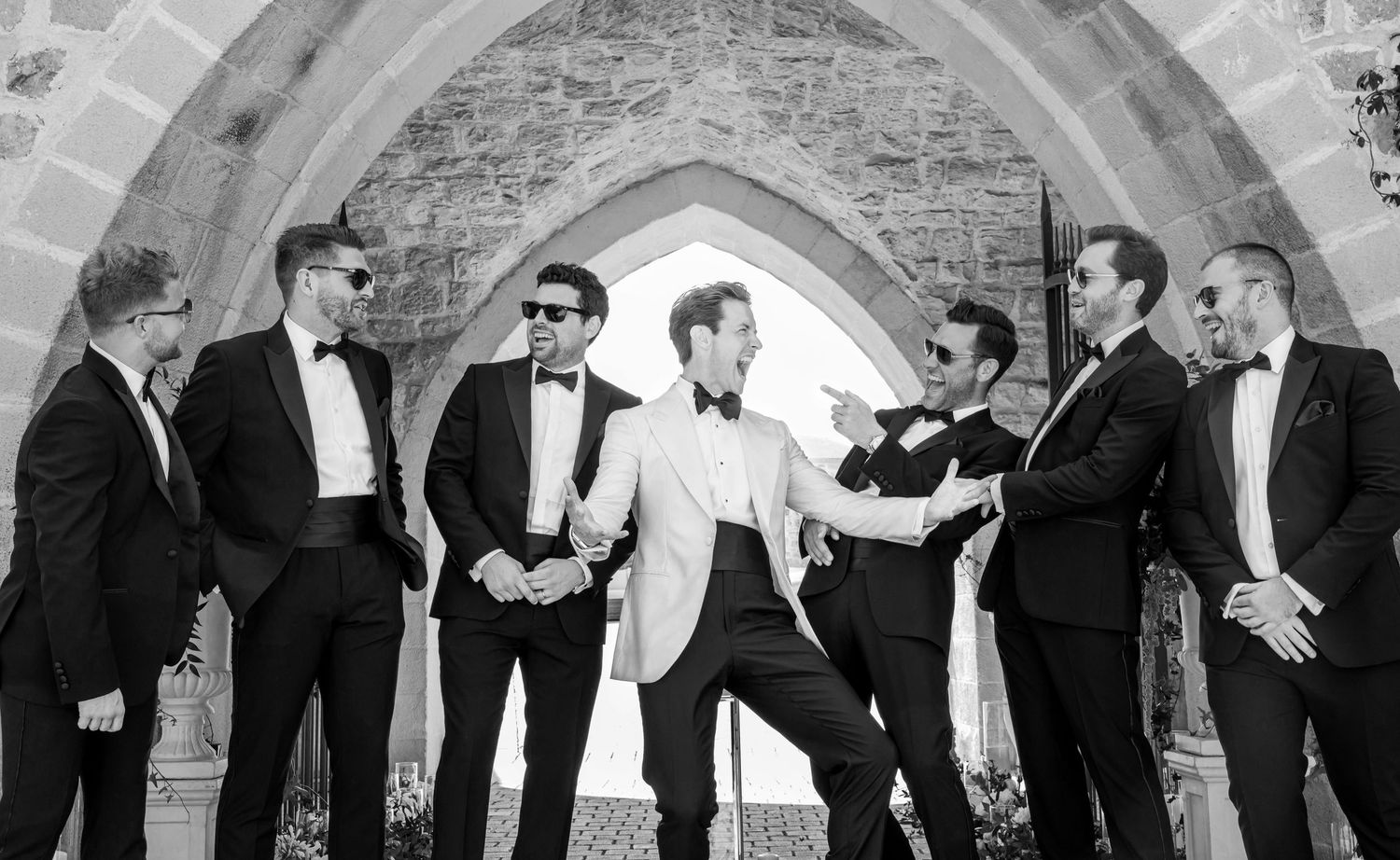 A black and white panoramic photo showing wedding guests dancing and celebrating in an elegant arched venue.