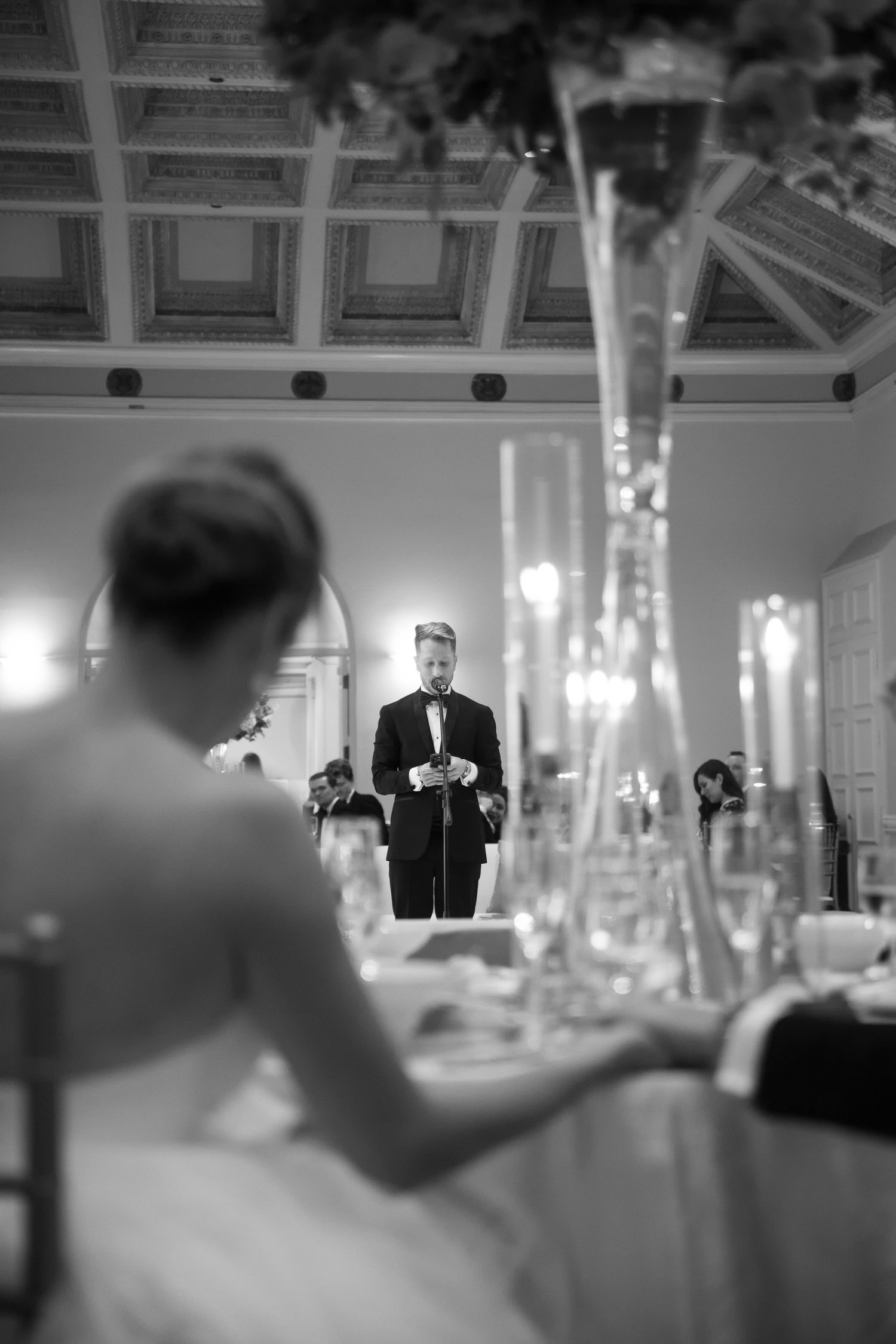 Black and white wedding reception photo of groom in tuxedo giving speech while bride sits in foreground.